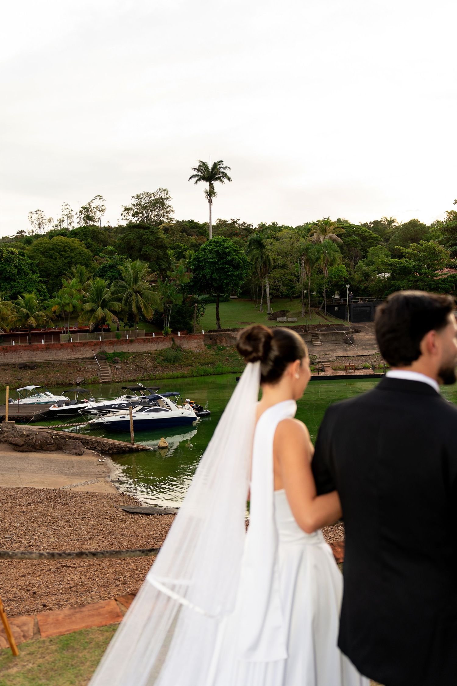 Bride in white gown with long veil and groom in tuxedo overlooking a tropical waterfront with boats and lush greenery.