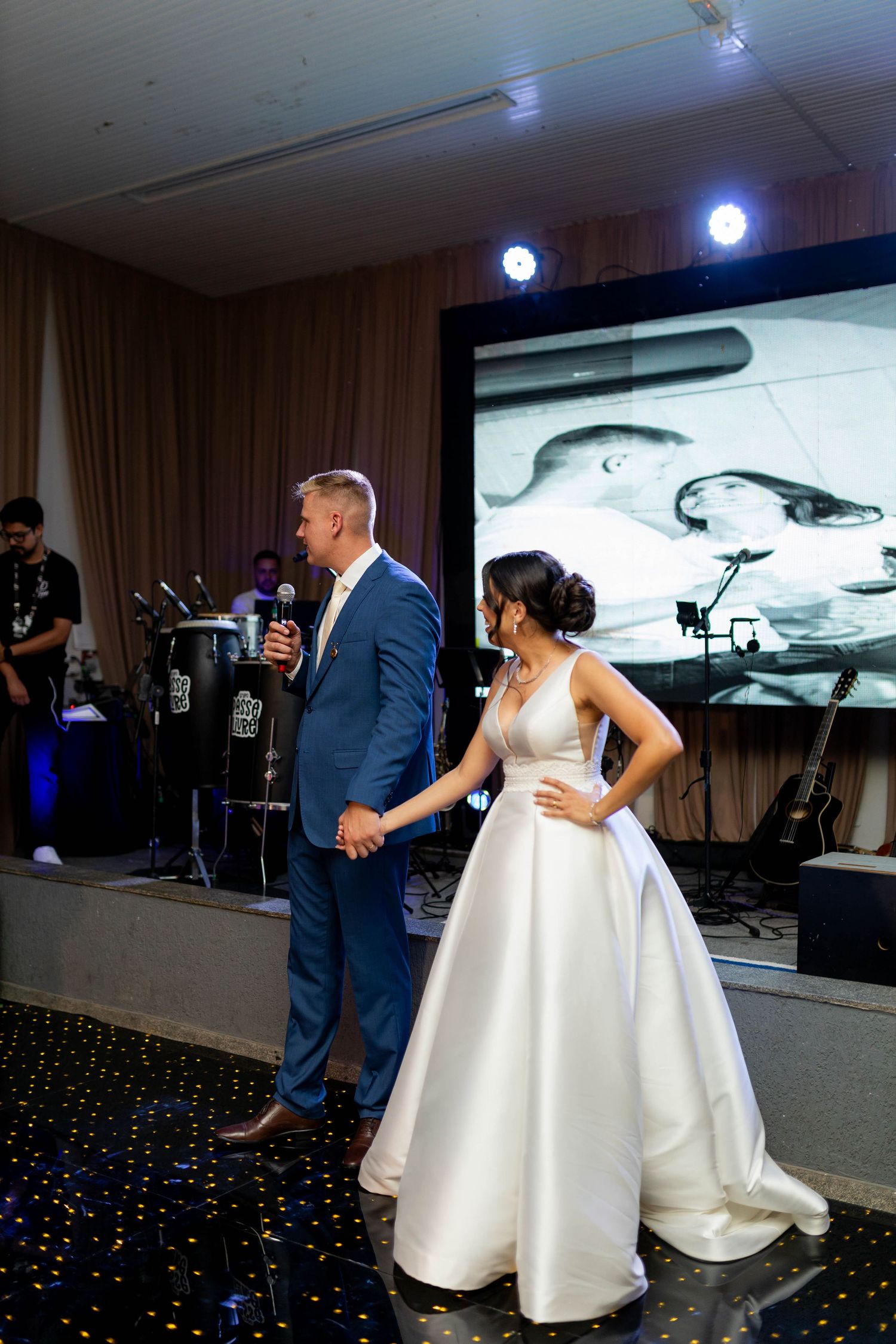 Bride in white gown and groom in blue suit holding hands on stage at elegant wedding reception.