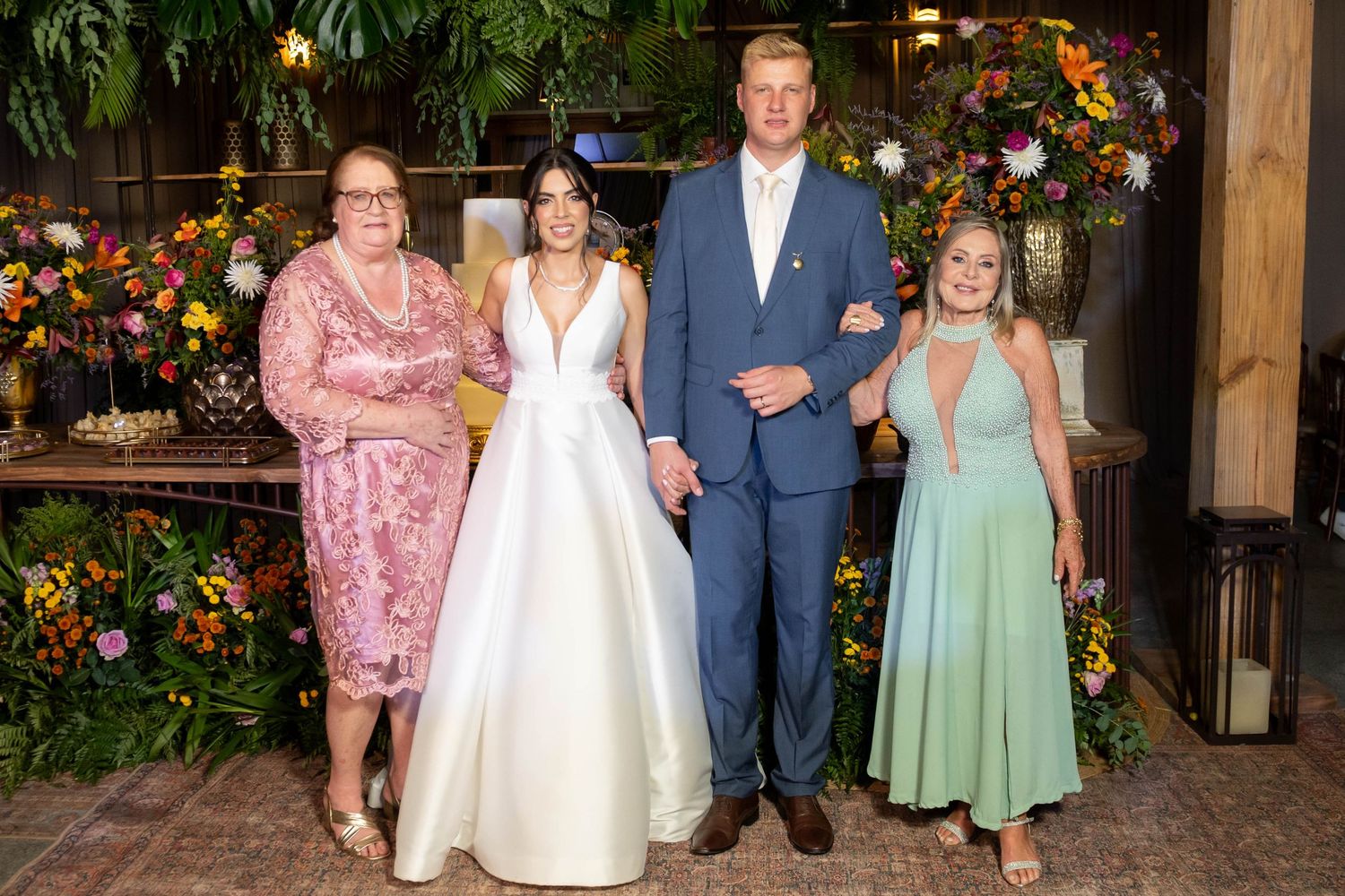 Wedding party of four posing before floral backdrop, bride in white gown, groom in blue suit, guests in pink and green dresses.