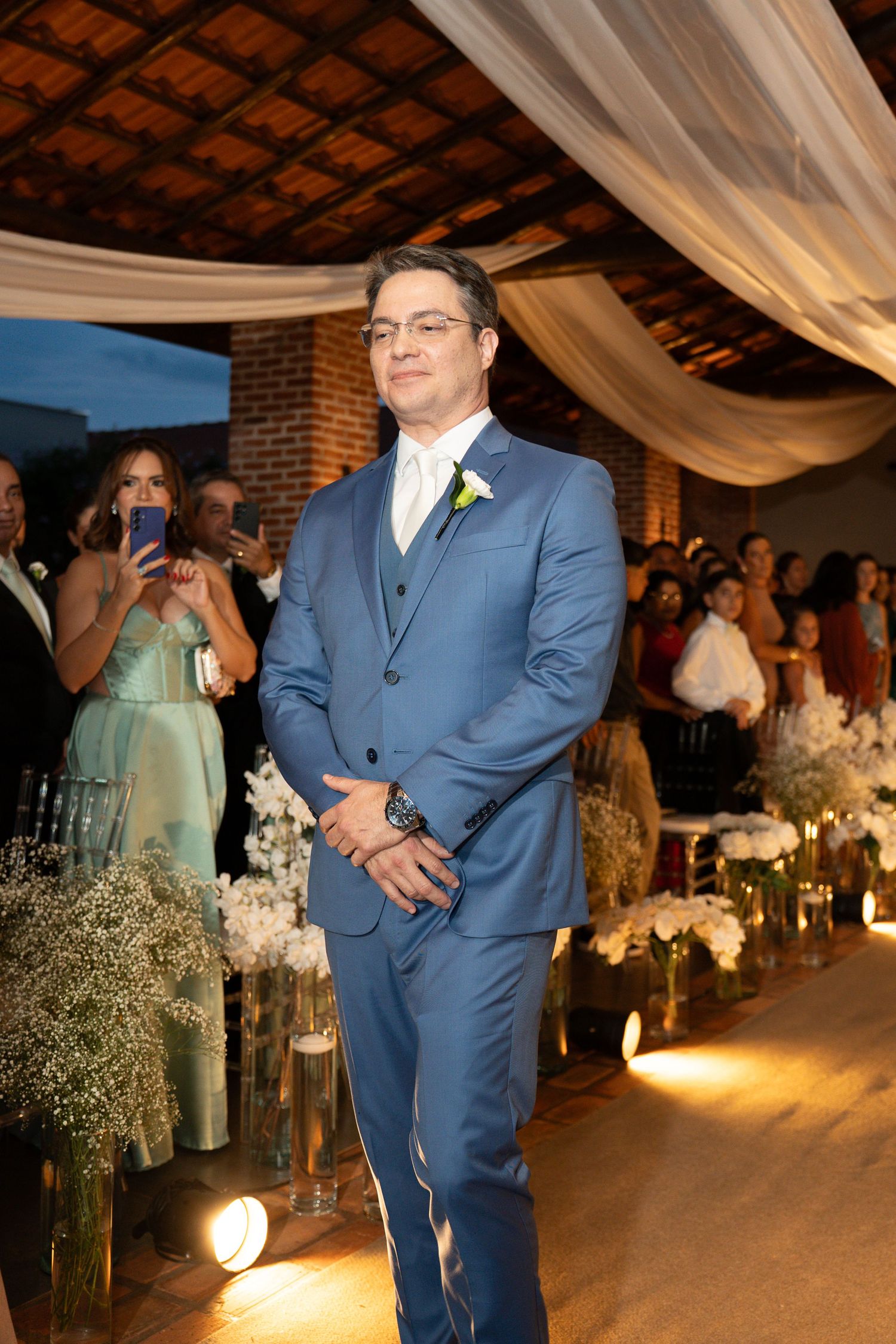 A groom in a blue three-piece suit with a white boutonniere stands at the altar during a wedding ceremony.