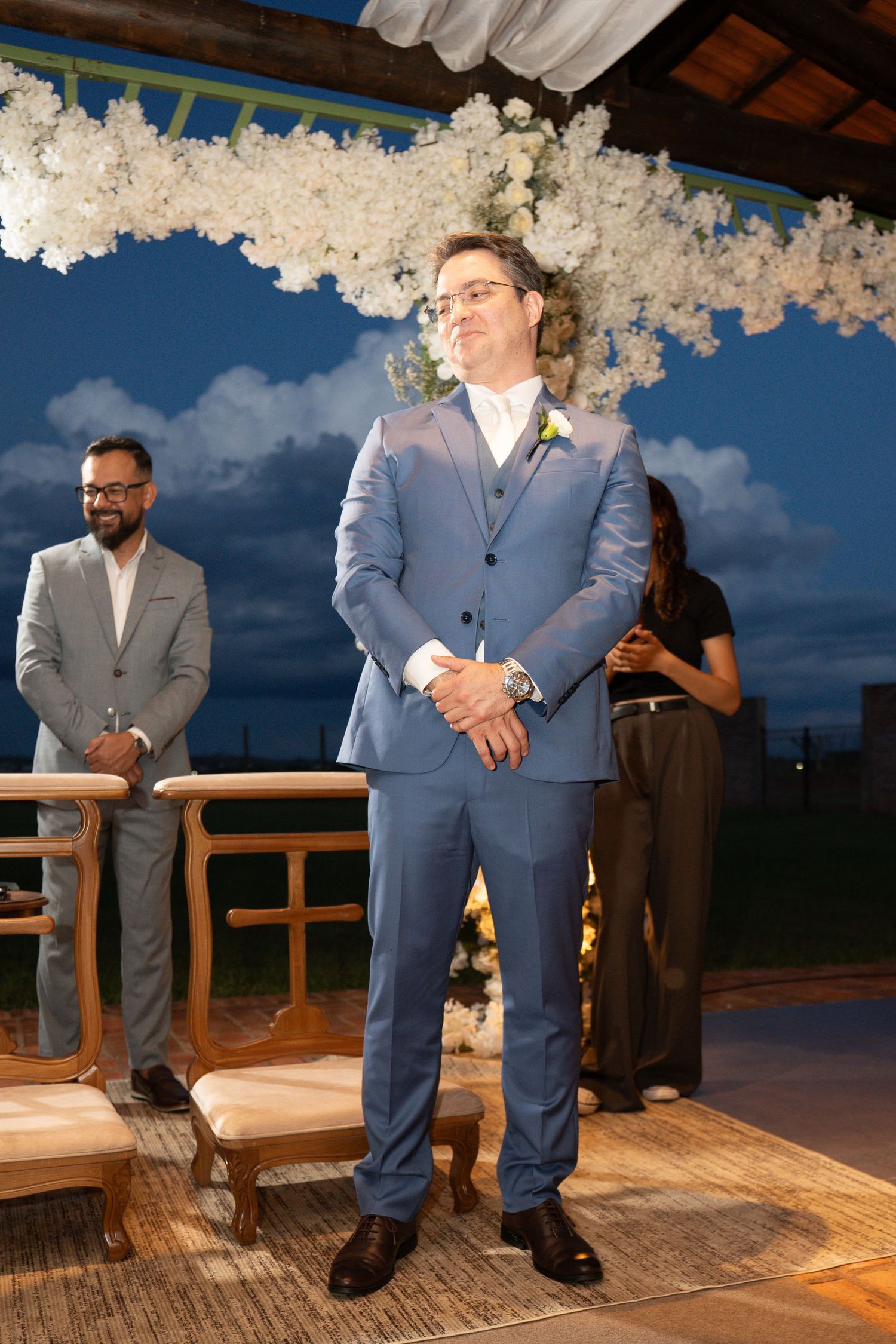 Groom in blue suit with boutonniere smiling at outdoor wedding ceremony with white floral arch at dusk.