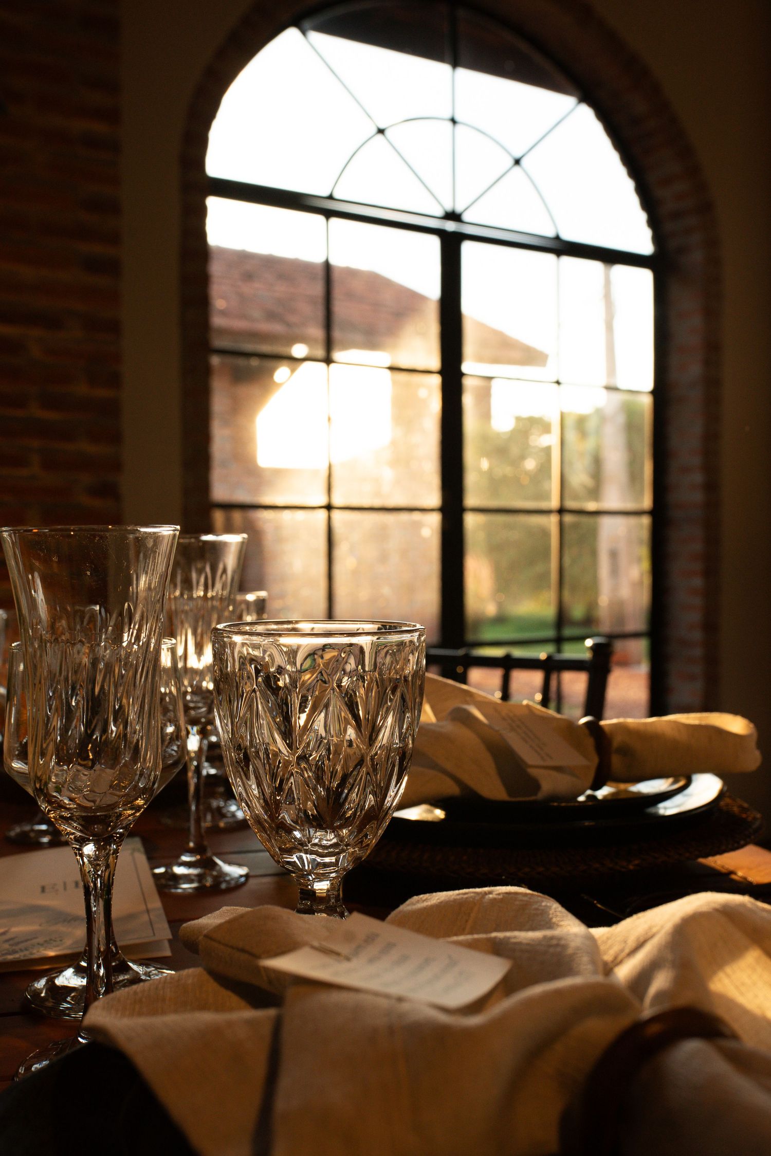 Crystal glassware and linen napkins on a dining table bathed in warm golden sunset light through an arched window.