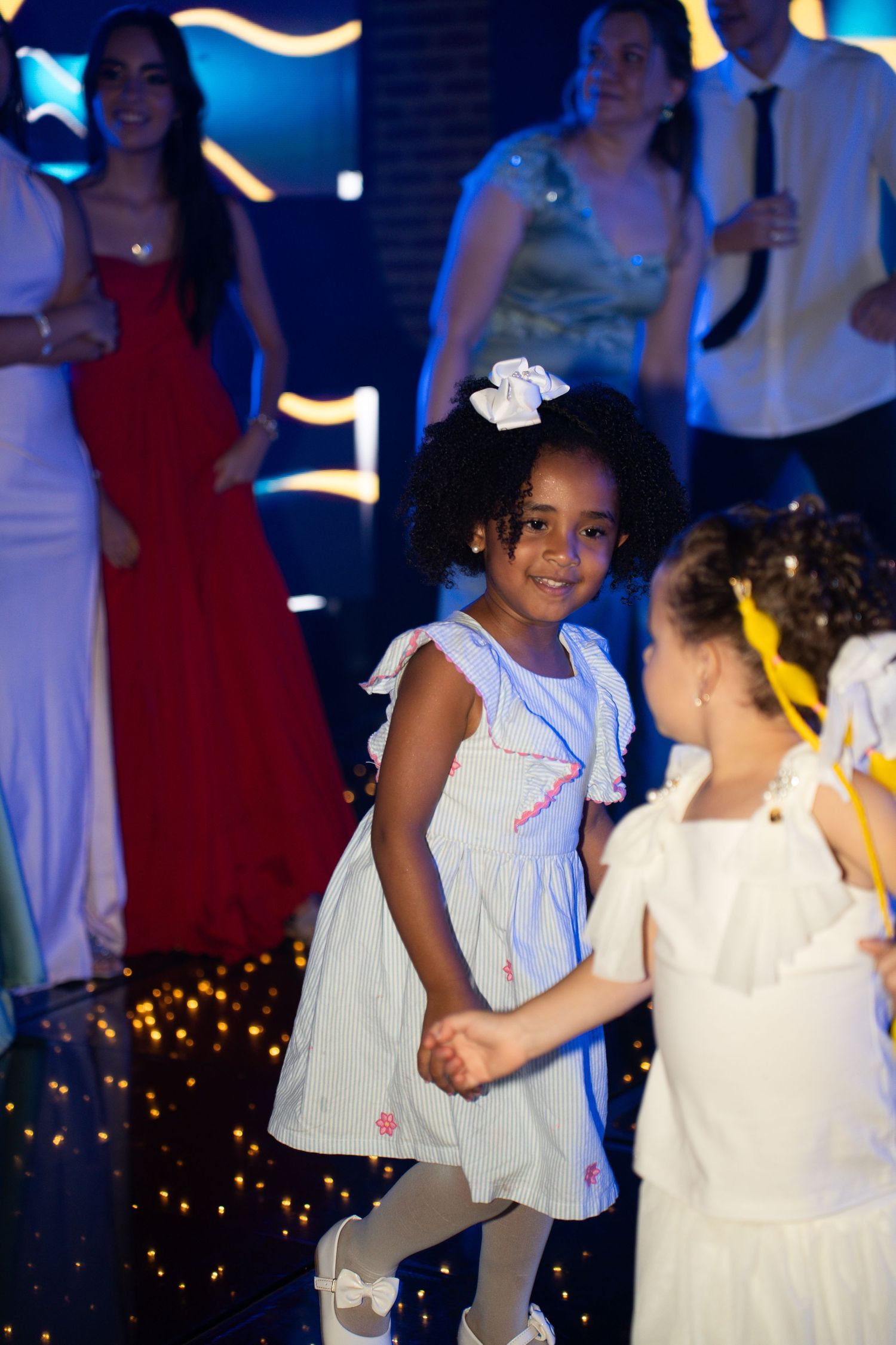 Two young girls in white dresses dancing together at a festive party with colorful lights in the background.