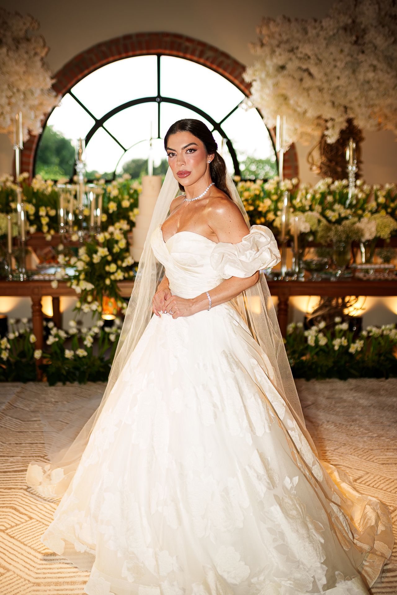 Bride in strapless white ballgown with veil poses elegantly before floral backdrop at luxurious wedding reception.