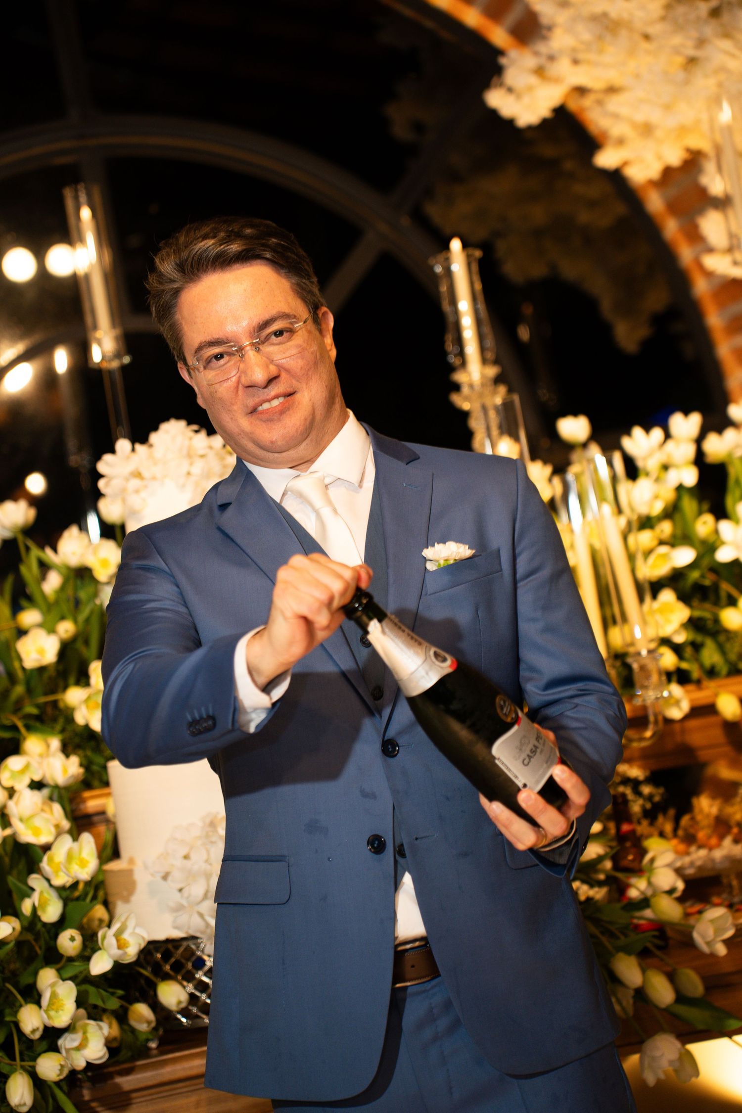 Elegant man in blue suit opening champagne bottle surrounded by white floral arrangements at formal event.