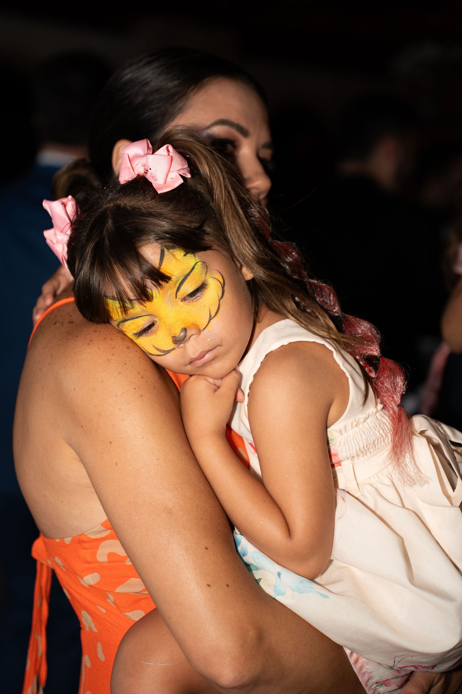 Tired little girl with tiger face paint resting on a woman's shoulder at a party.