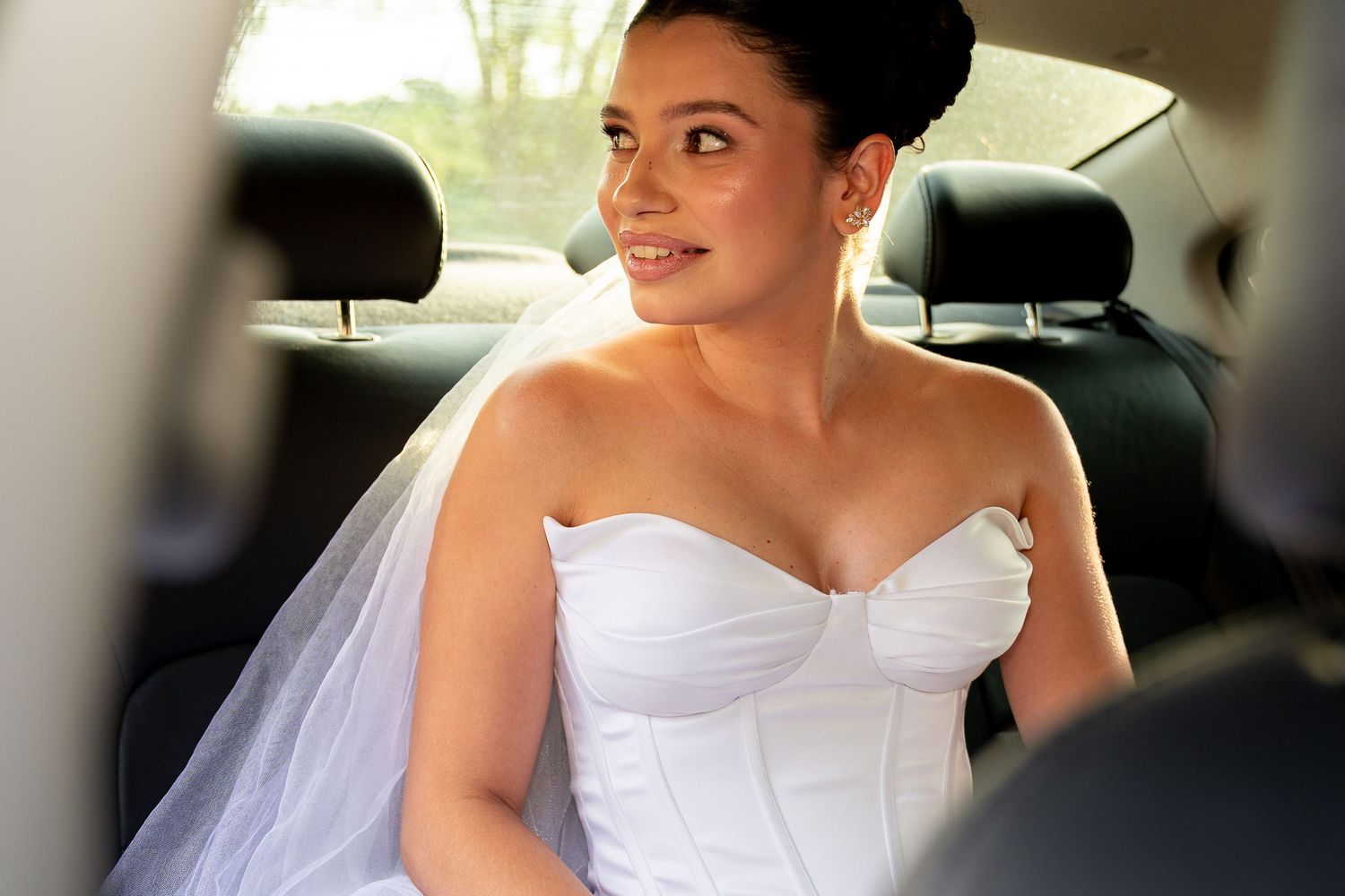 A beautiful bride in a white strapless gown and veil sits in a car, smiling elegantly on her wedding day.