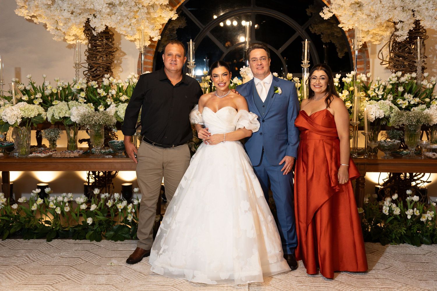 Wedding party of four posing elegantly, bride in white gown, groom in suit, guests in formal attire, floral decor background.