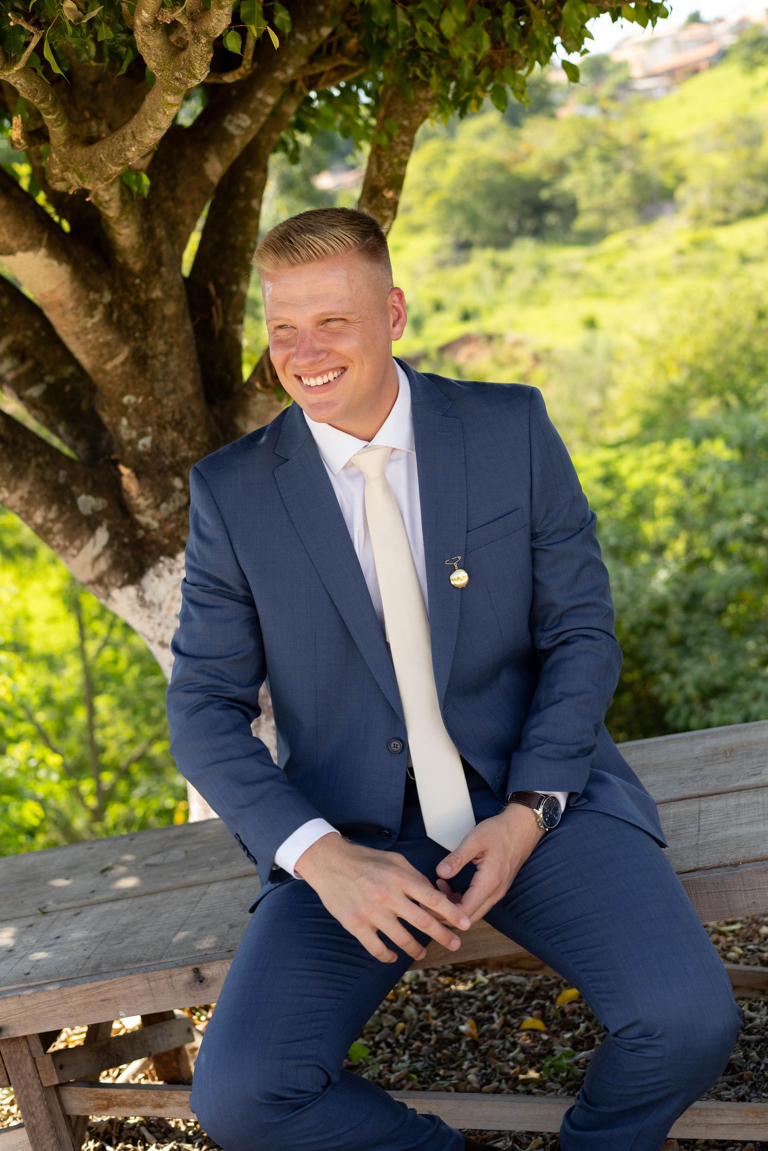 Smiling young man in navy blue suit and cream tie sitting outdoors under a tree with lush green hills behind him.