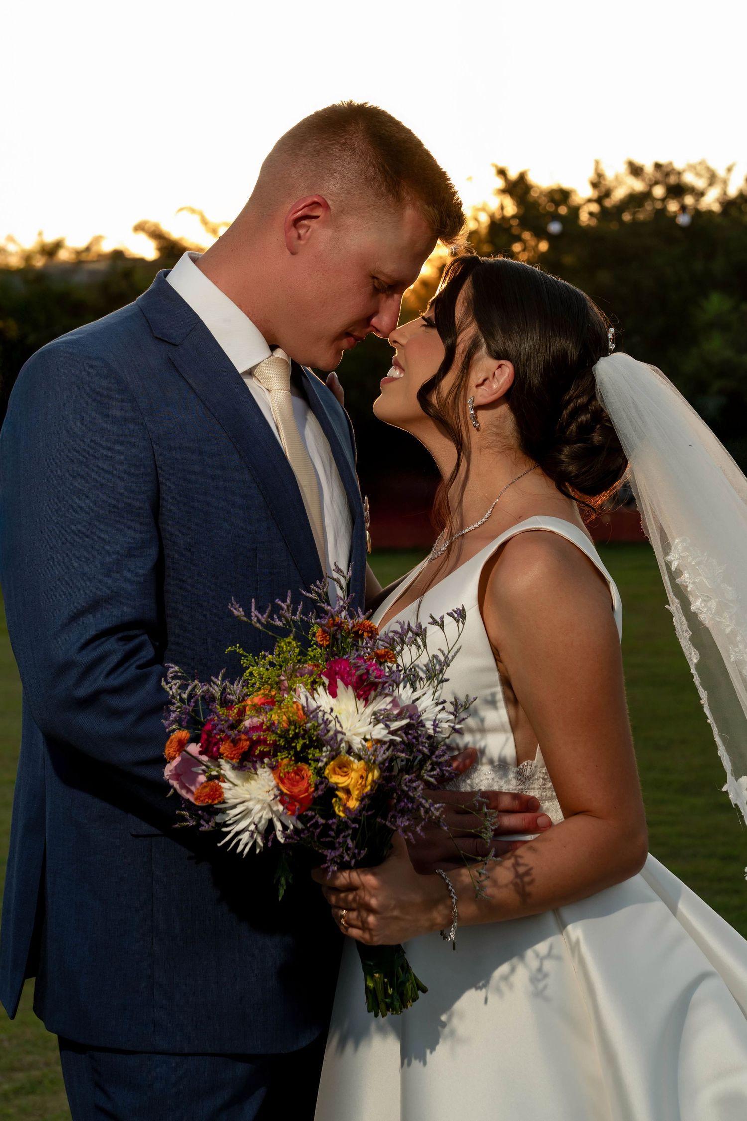 Bride and groom share a romantic moment at golden hour, she holds a colorful bouquet while wearing a veil and white gown.