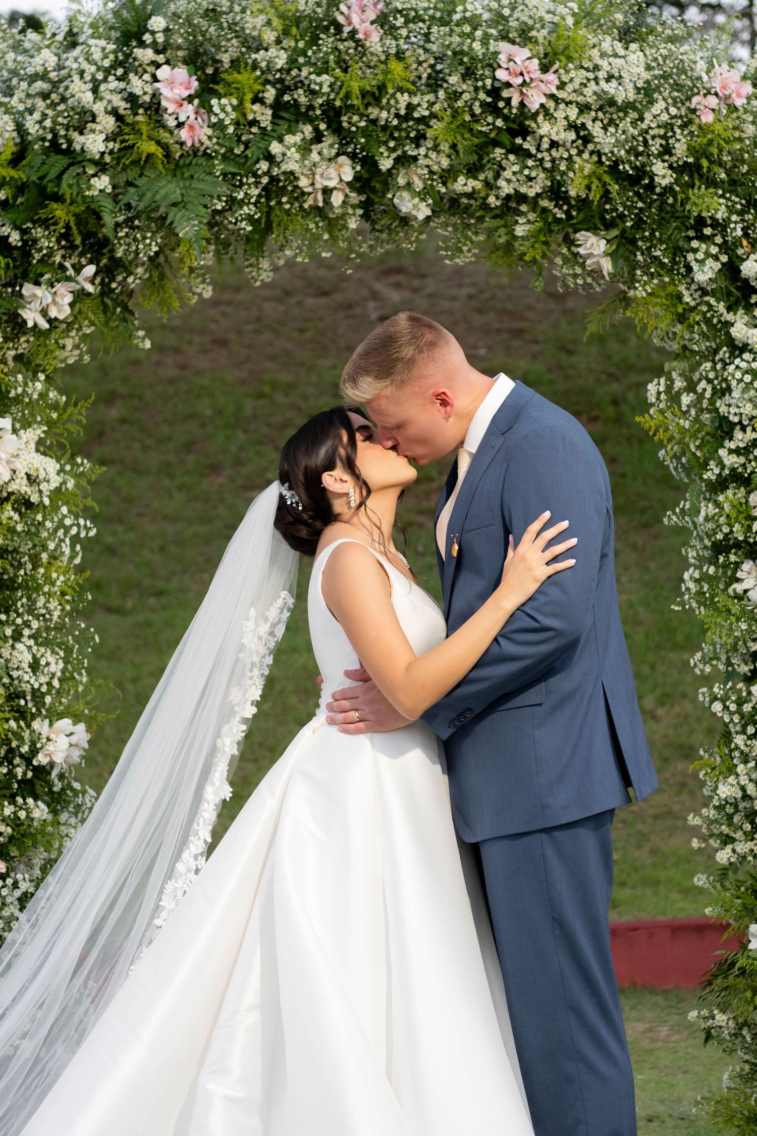 Bride and groom share a romantic kiss under a floral arch adorned with white and pink flowers at an outdoor wedding.