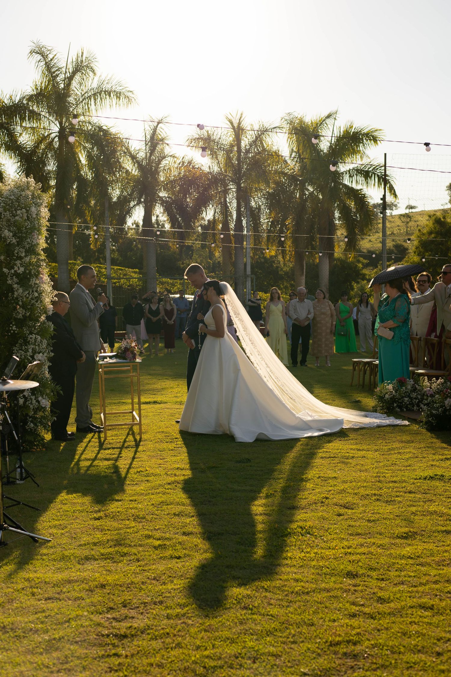 Bride and groom share a kiss during an outdoor wedding ceremony on lush green grass surrounded by palm trees.