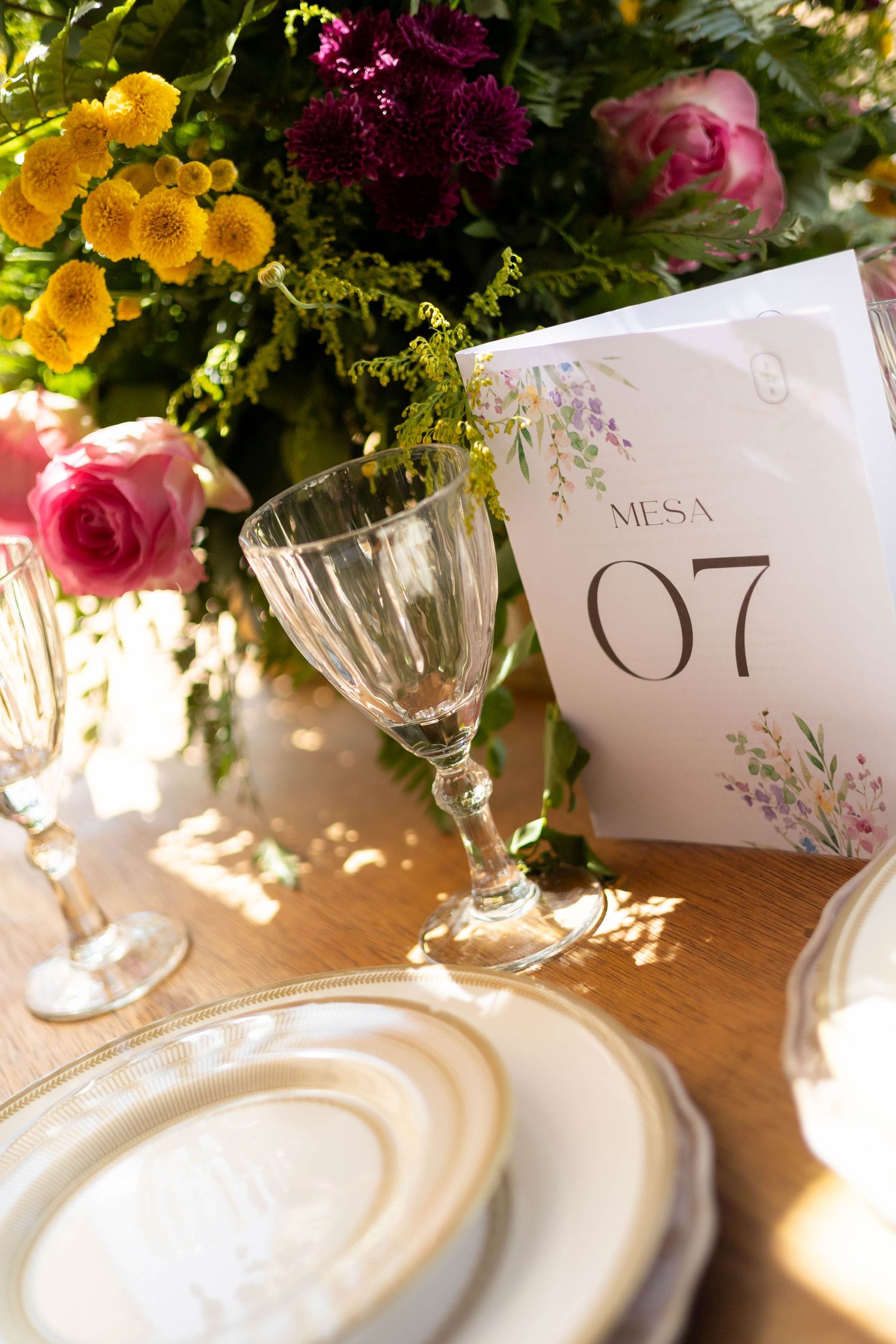 Elegant wedding table setting with crystal glasses, gold-rimmed plates, colorful flowers, and a Mesa 07 table number card.