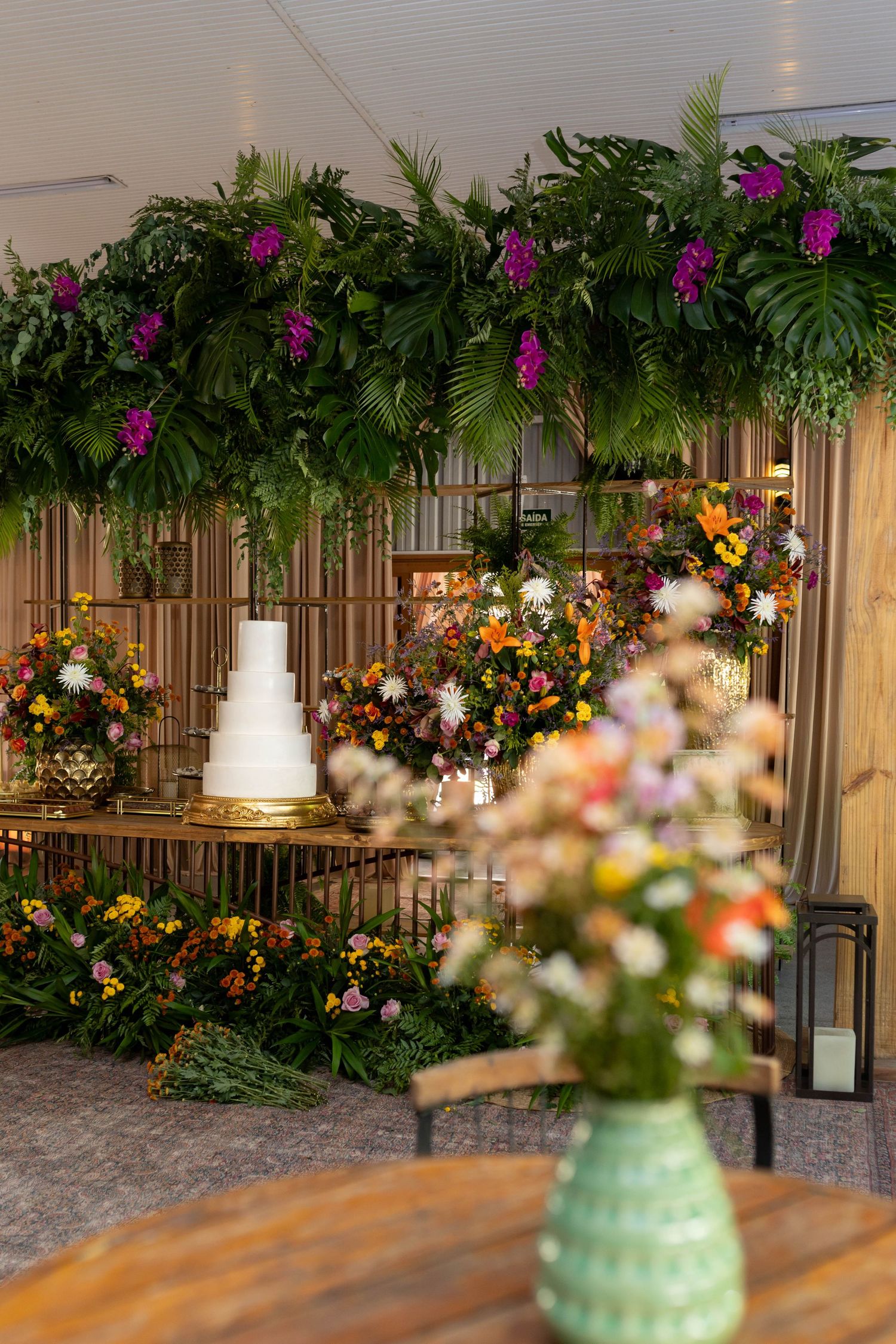 Elegant wedding cake display surrounded by lush tropical greenery and colorful floral arrangements under a decorated canopy.
