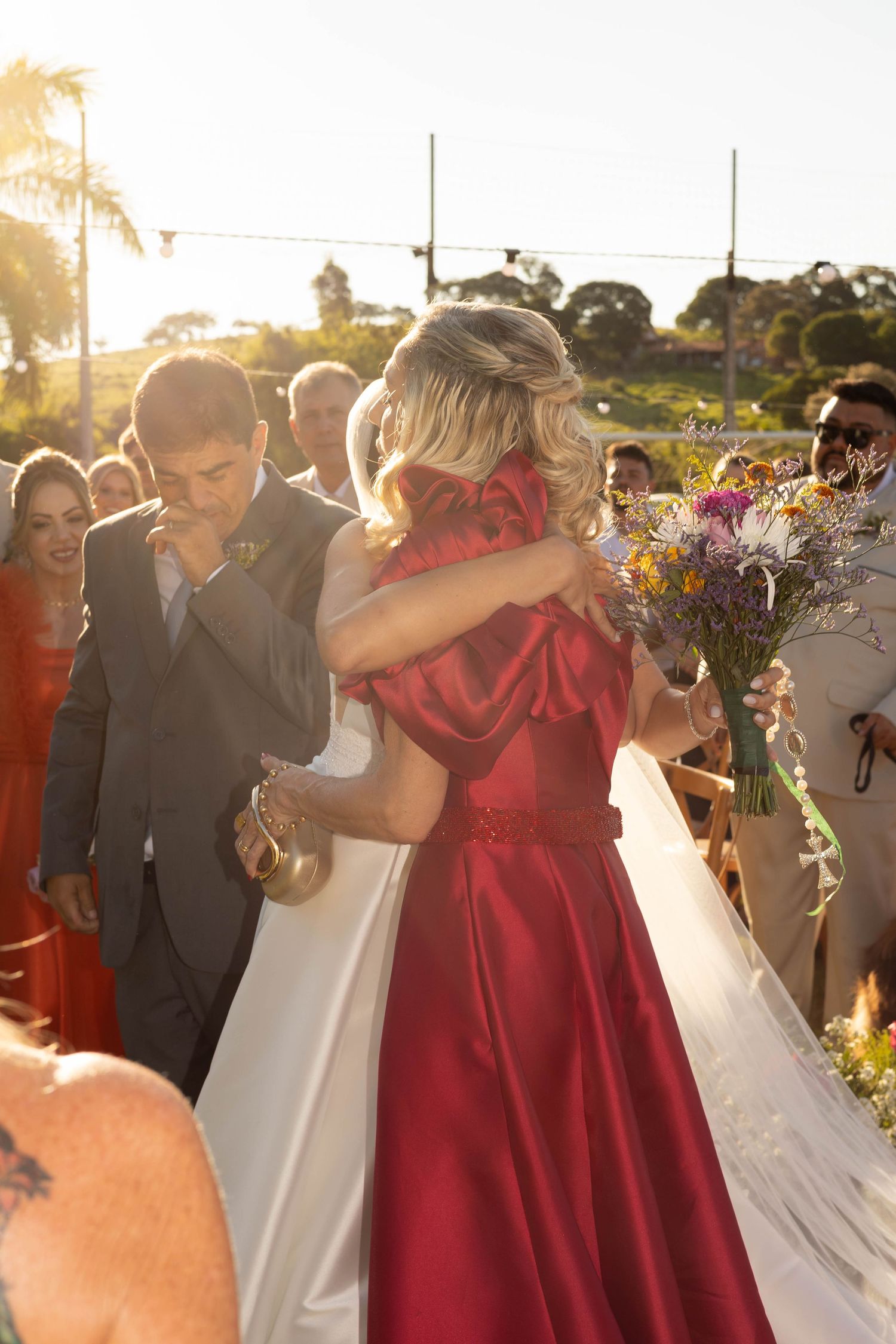 Bride in white gown embraces woman in red satin dress during outdoor wedding ceremony at golden hour.