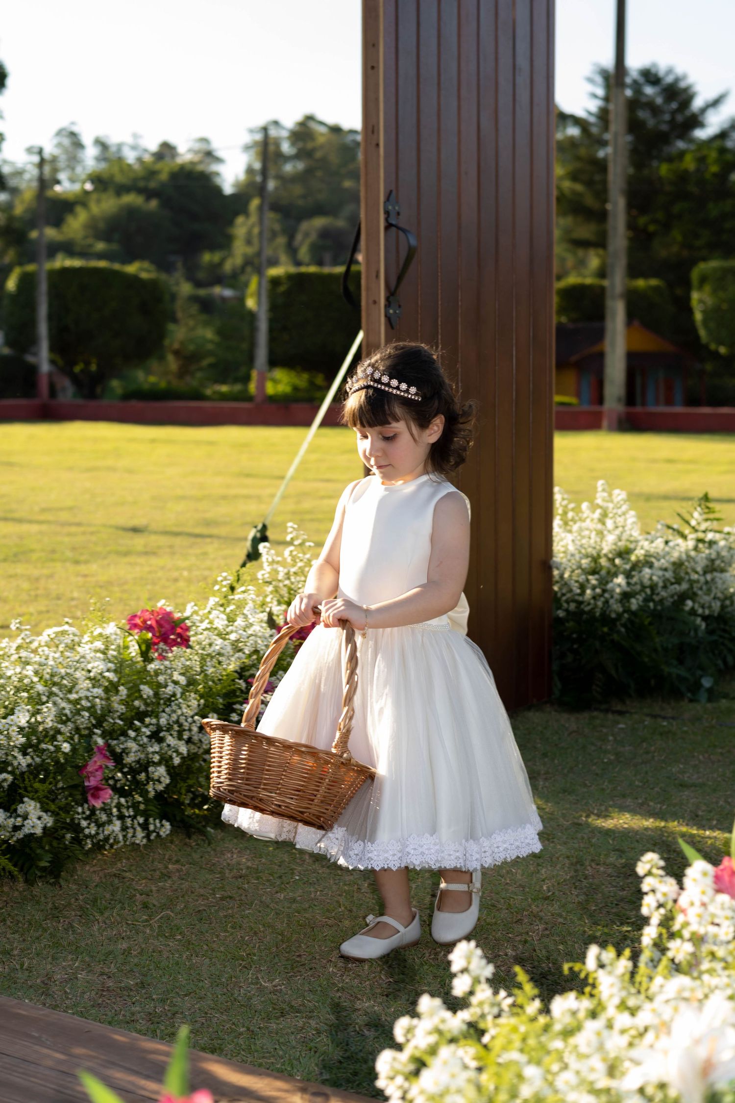 Young flower girl in white dress holding wicker basket among garden flowers at outdoor wedding ceremony.