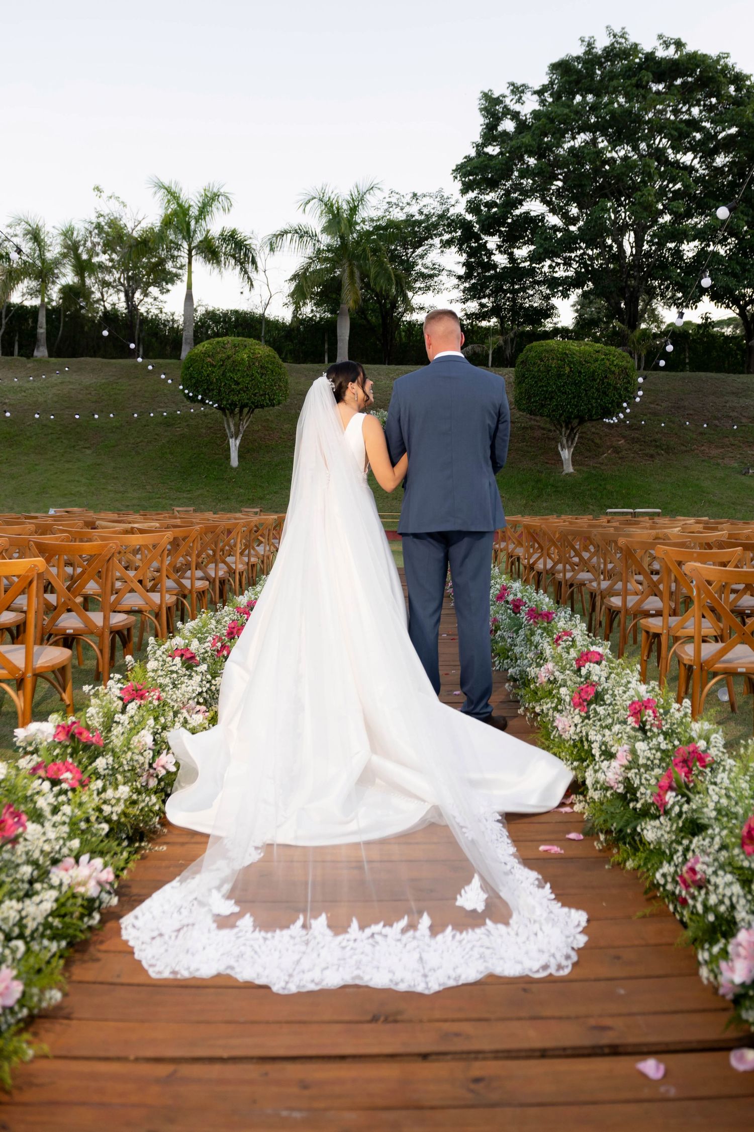 Bride and groom walking down floral-lined aisle at outdoor wedding ceremony with lace cathedral veil trailing behind.