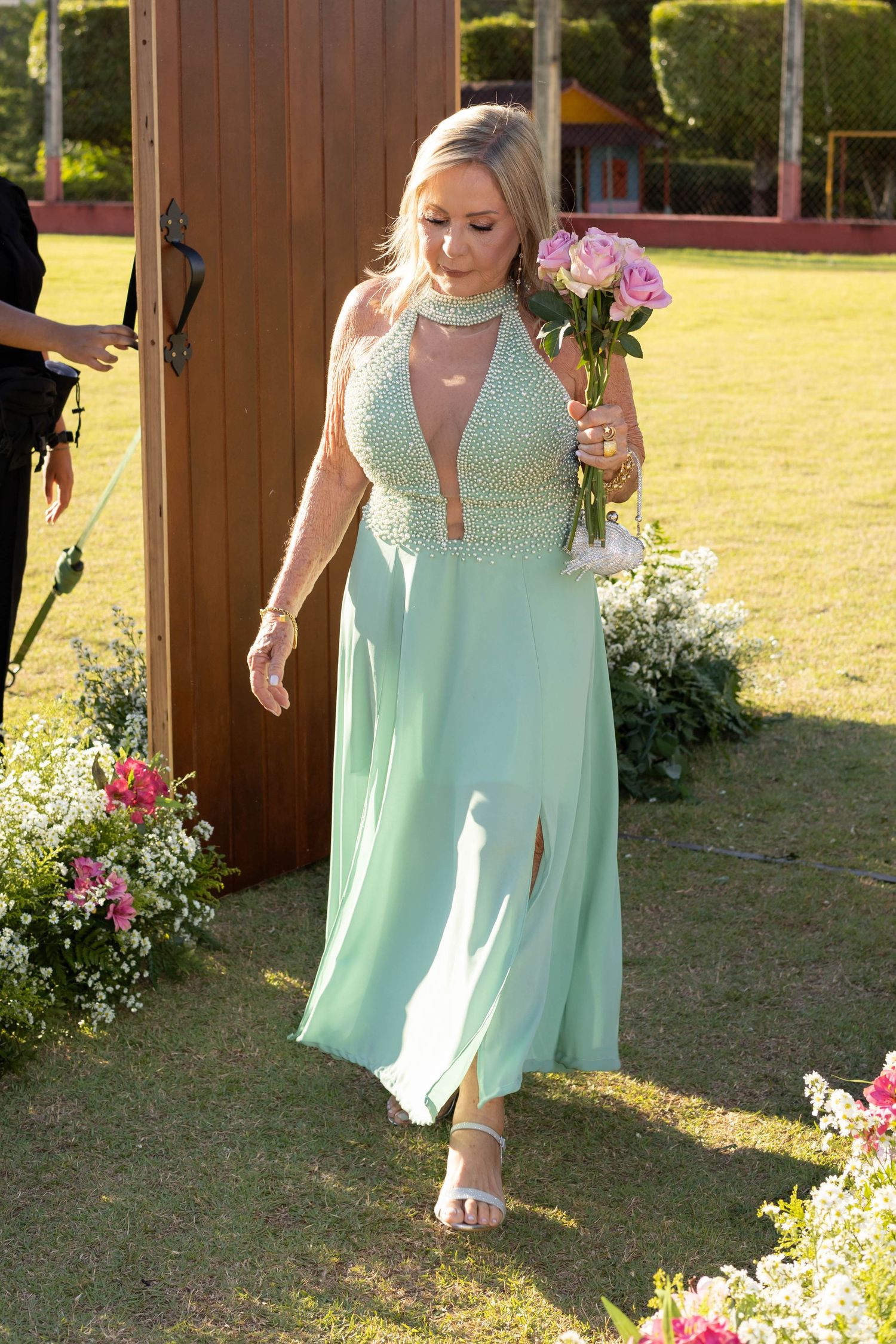 Woman in mint green halter gown holding pink roses walks through floral garden at outdoor wedding.