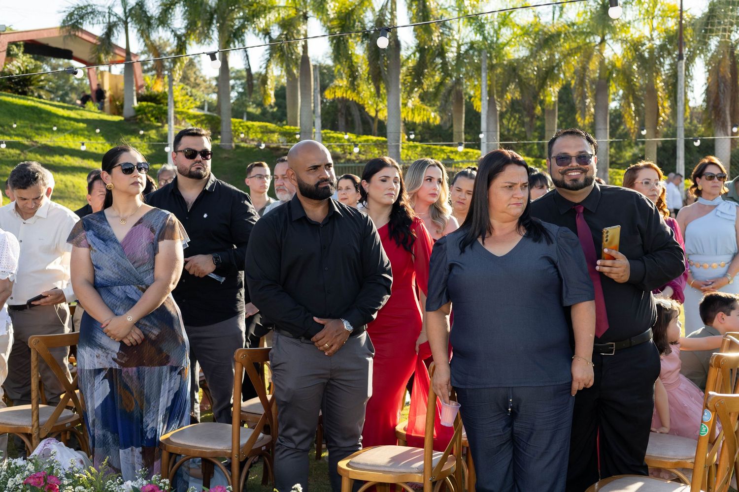 Guests standing at an outdoor wedding ceremony surrounded by lush palm trees and floral decorations.