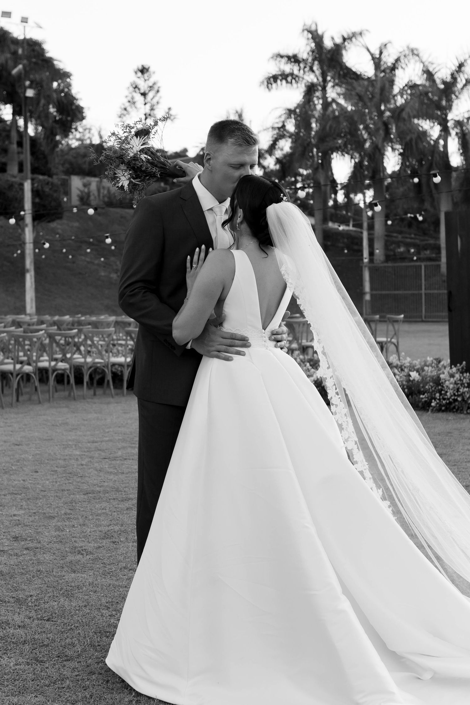 Black and white photo of a bride and groom sharing a romantic embrace outdoors with palm trees and wedding chairs in the background.