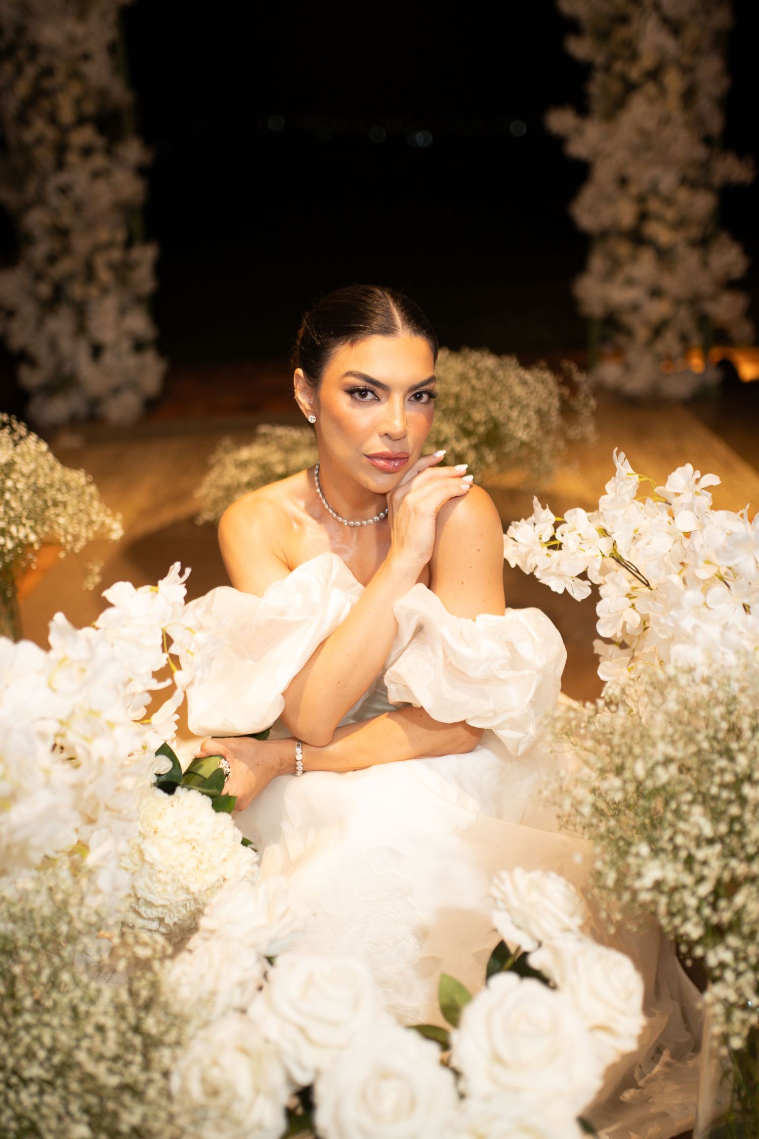 Elegant bride in white ruffled gown surrounded by white roses and floral arrangements in a romantic night setting.