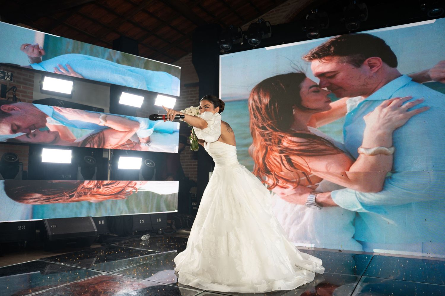 Bride in white gown poses before large screens displaying romantic couple photos at elegant wedding reception.