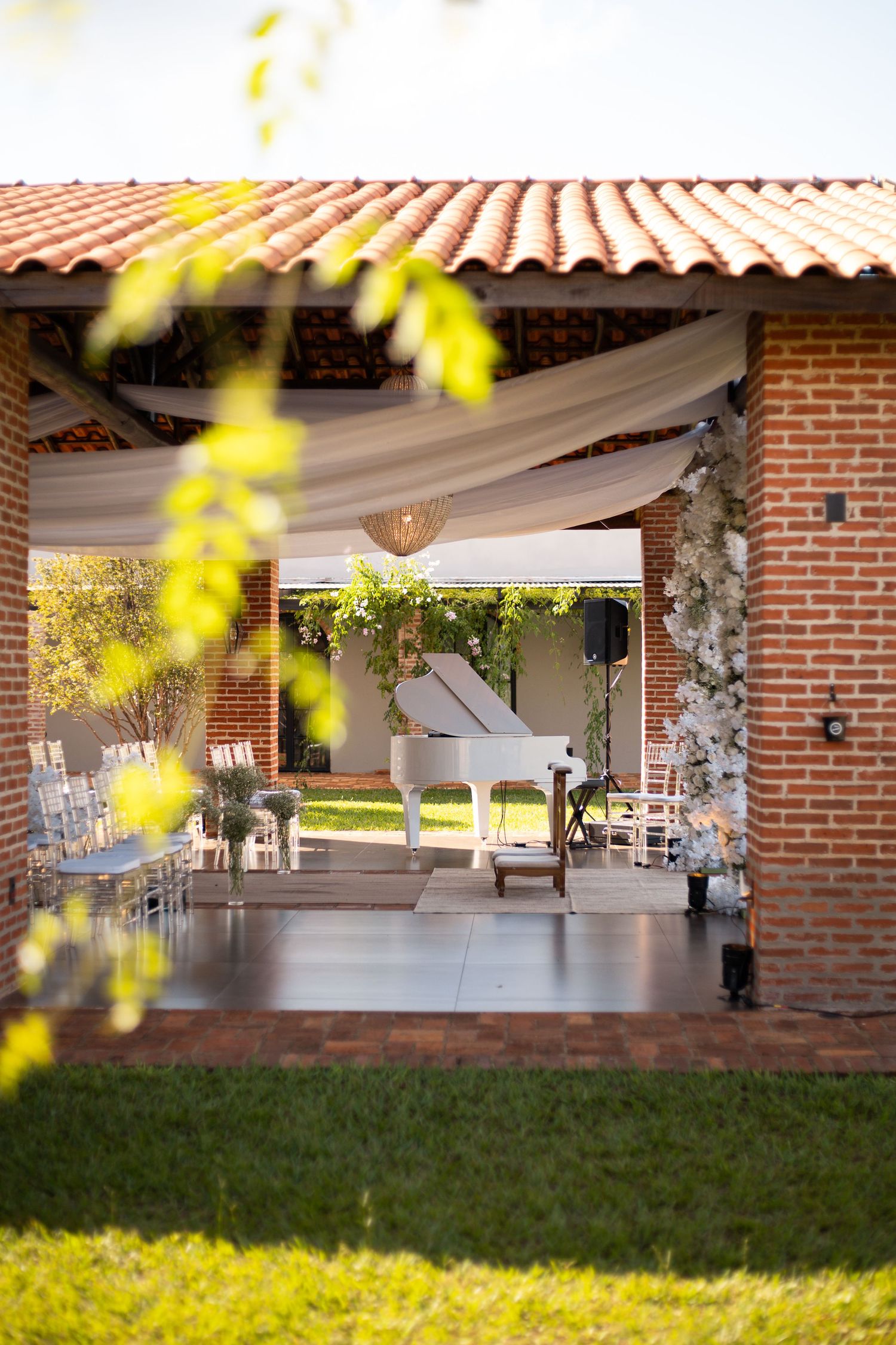 Elegant outdoor wedding venue with white grand piano, floral arch, draped fabric ceiling, and brick walls under terracotta roof.