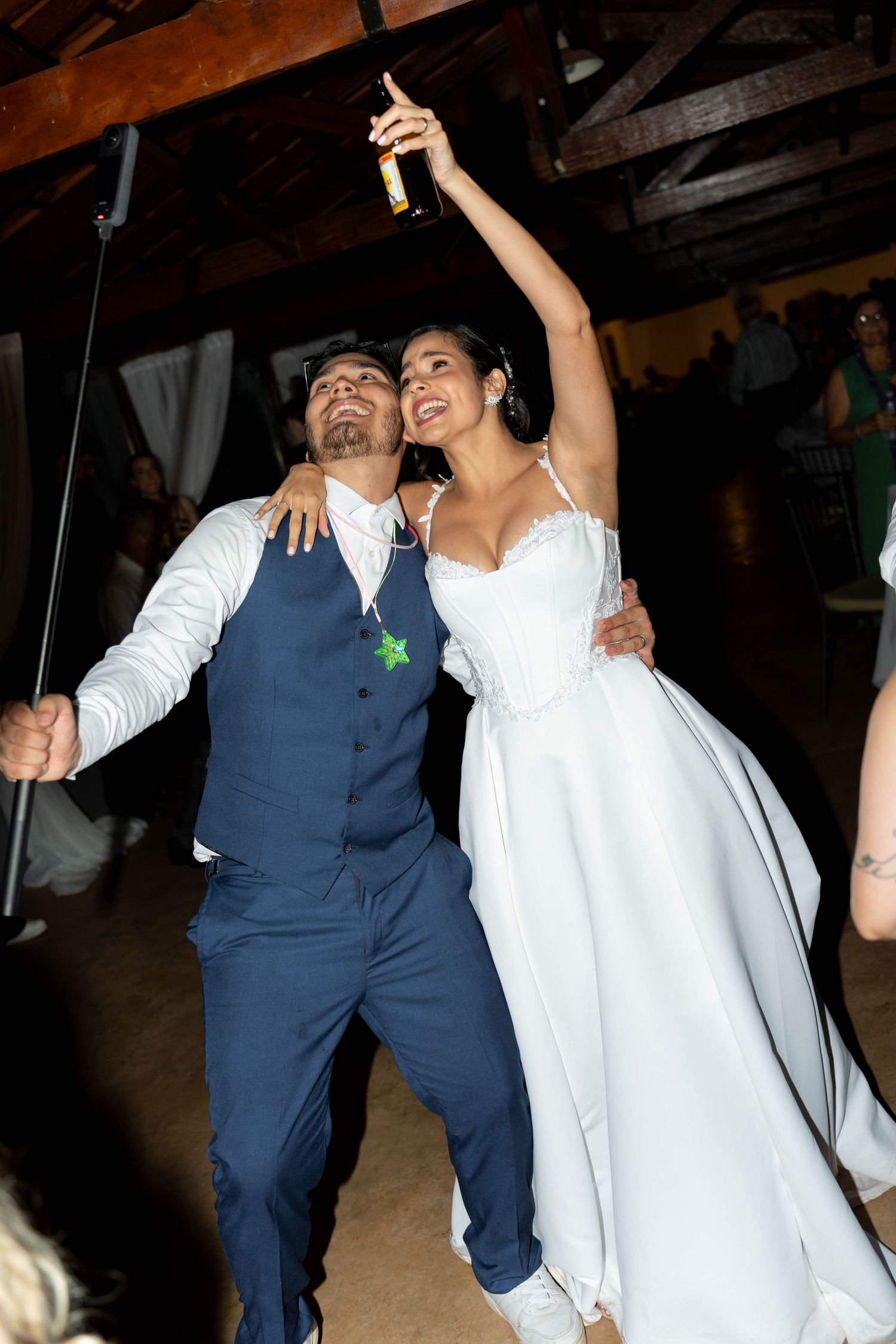 Joyful bride in white gown raises bottle while groom in navy suit embraces her on wedding reception dance floor.