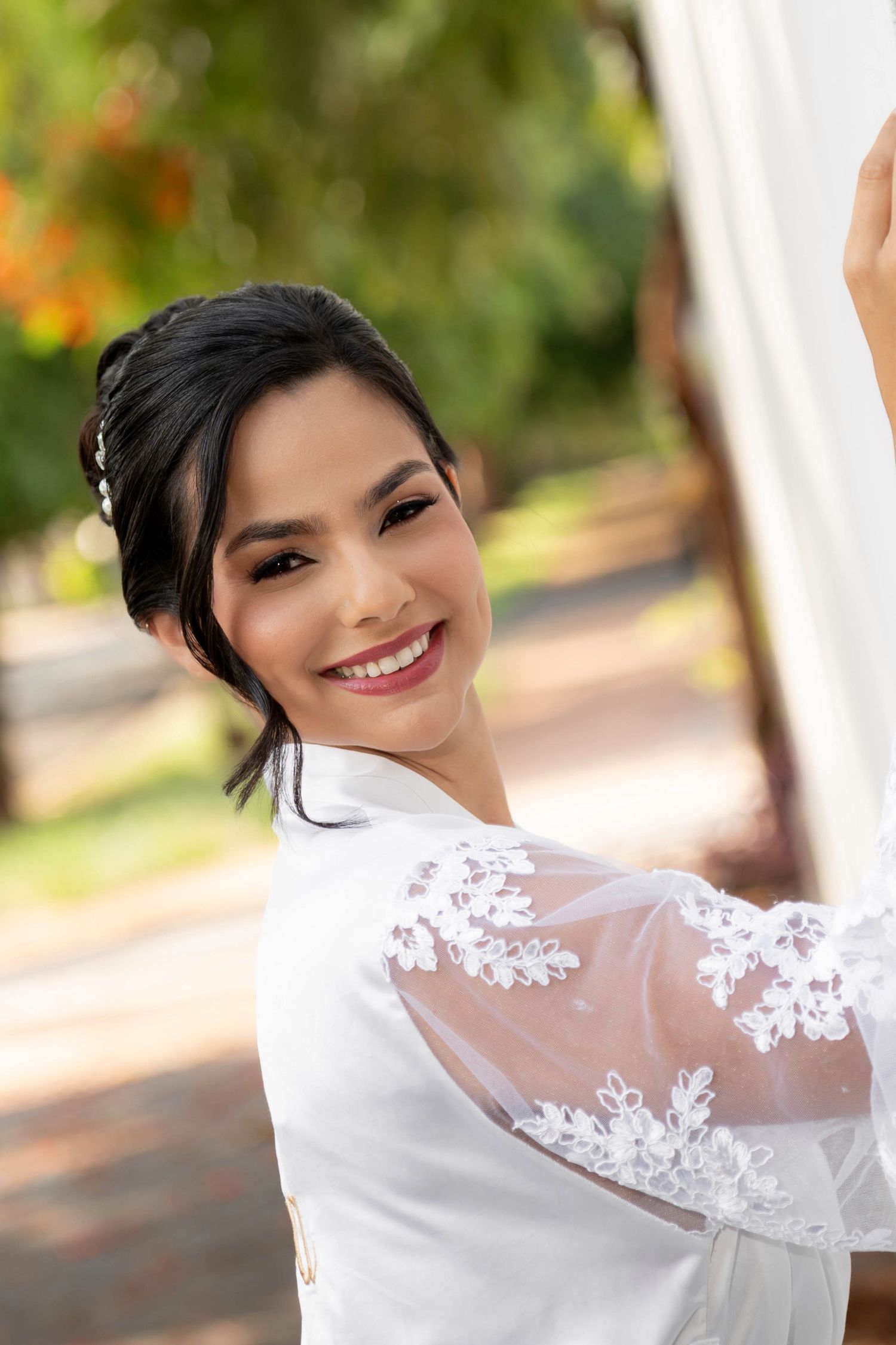 Smiling bride in white lace gown poses outdoors among green trees on her wedding day.