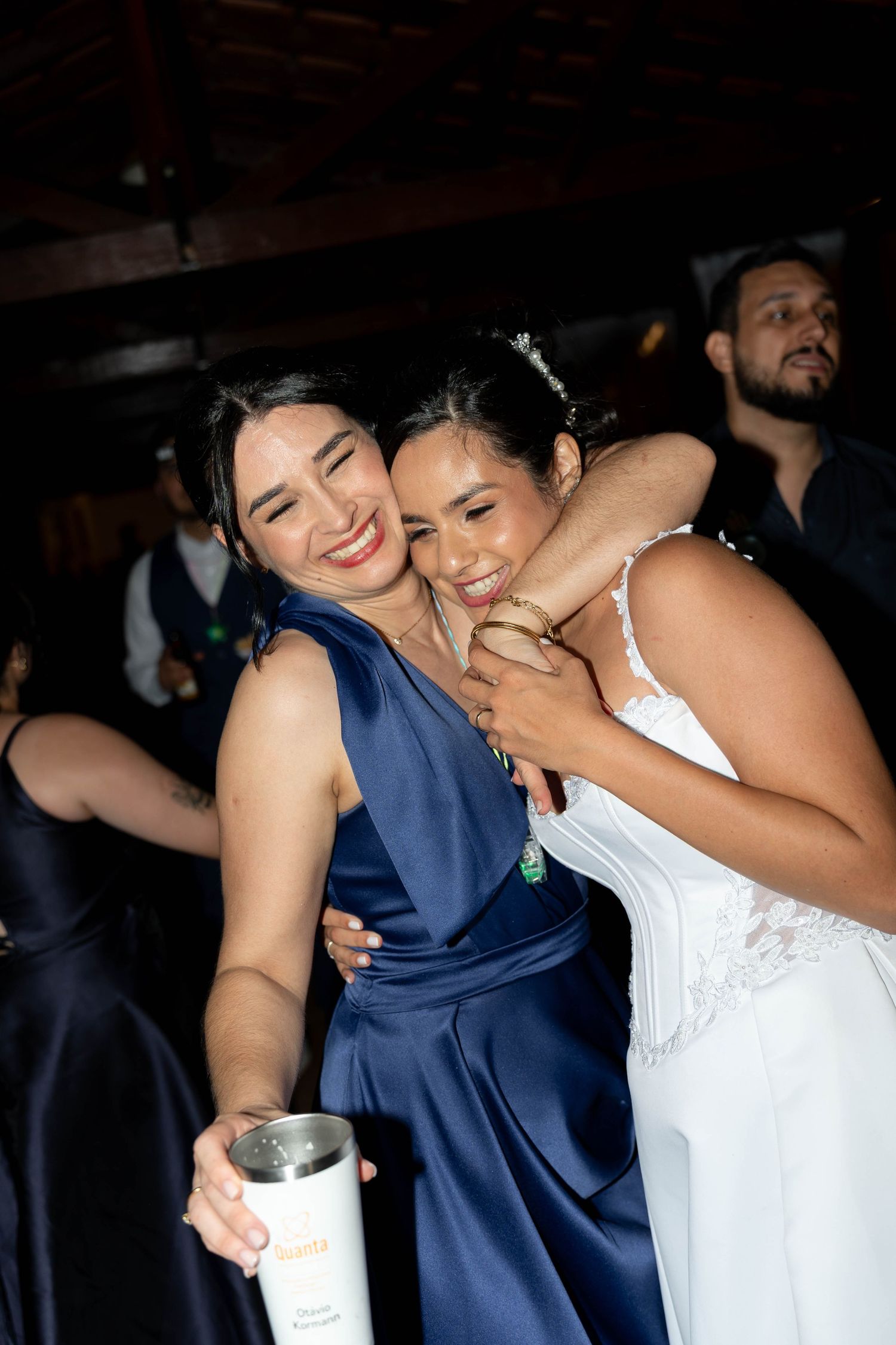 Bride in white gown hugs bridesmaid in navy blue dress at wedding reception celebration.
