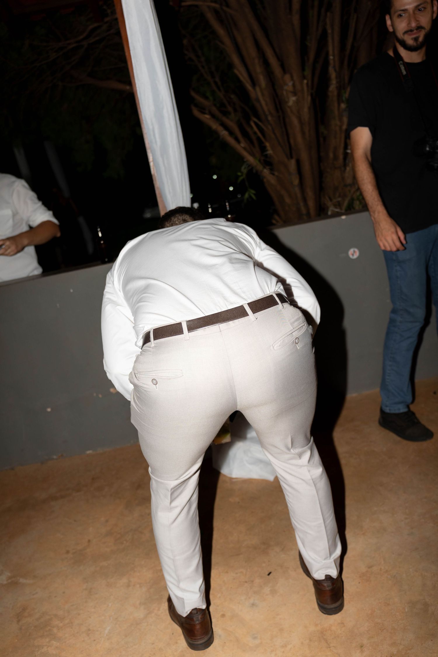 Man in white shirt and pants bending over at an outdoor evening event with guests nearby.