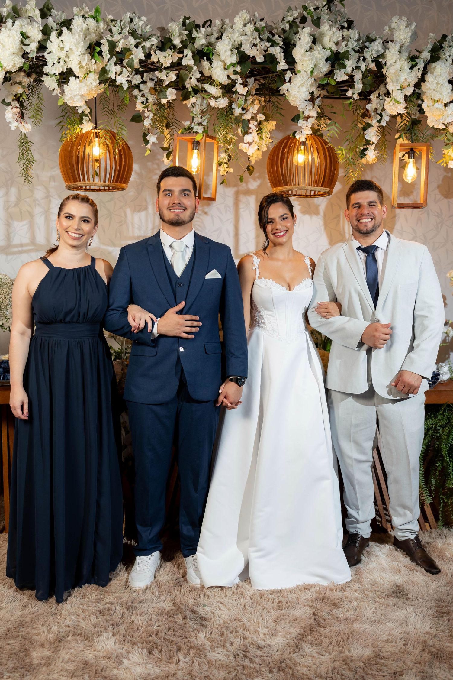 Wedding party of four posing elegantly under floral arch with warm pendant lights, bride in white gown with groom in navy suit.