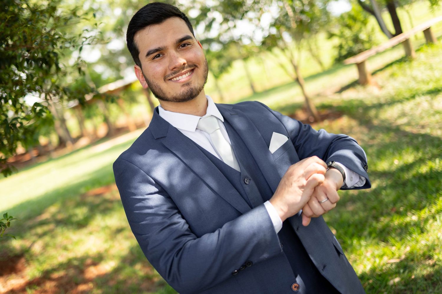 Smiling young man in a navy blue suit adjusting his cufflinks outdoors in a green park setting.