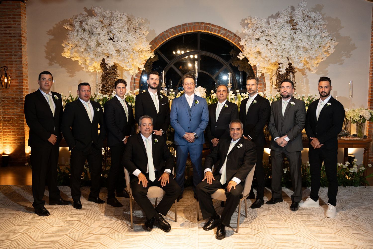 Groom in blue suit poses with groomsmen in black tuxedos at elegant wedding venue with floral arch backdrop.