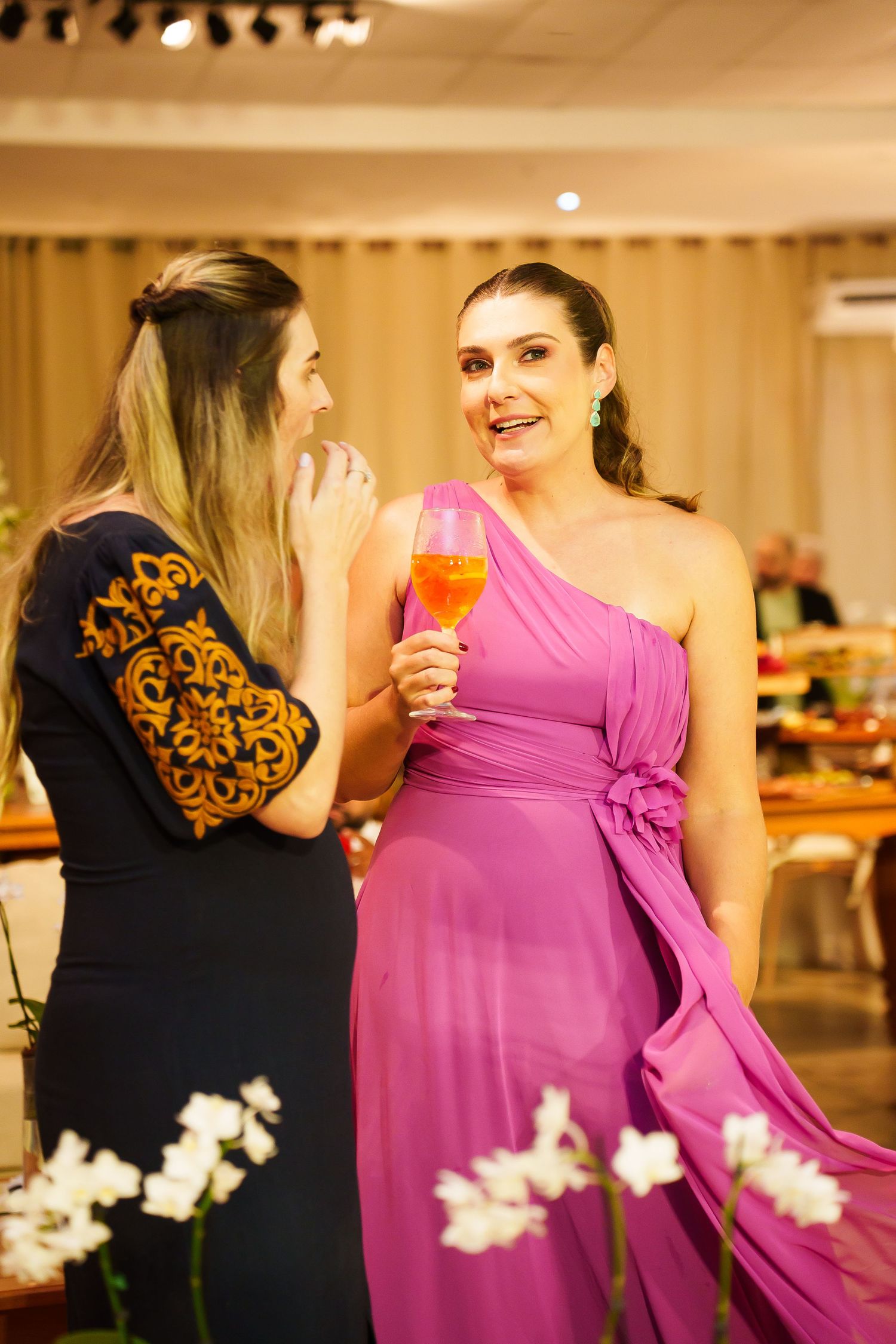 Two women in elegant dresses chat at a party, one in pink holding a cocktail glass.