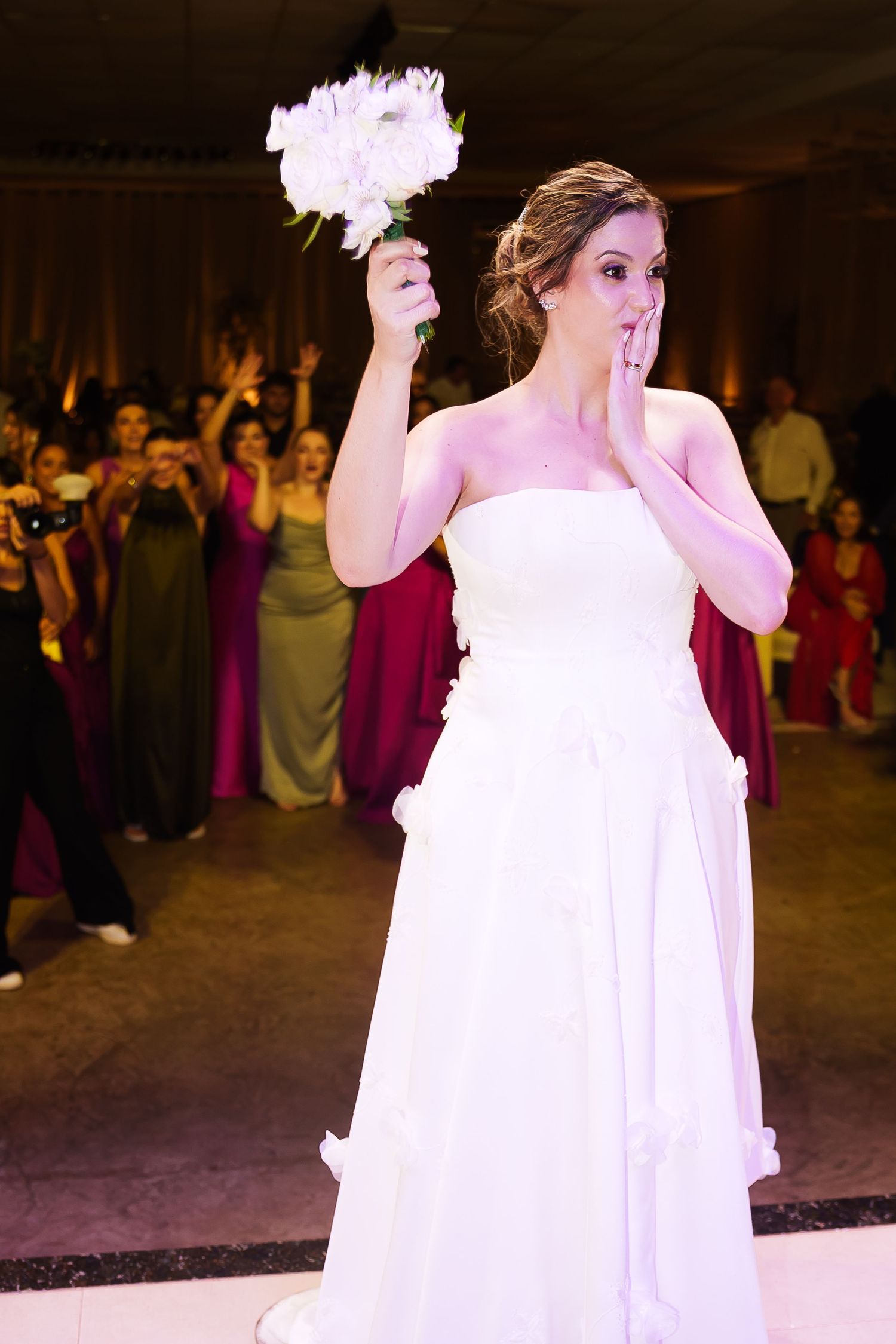 Bride in white strapless gown raises bouquet before toss, hand over mouth, bridesmaids eagerly waiting behind her.