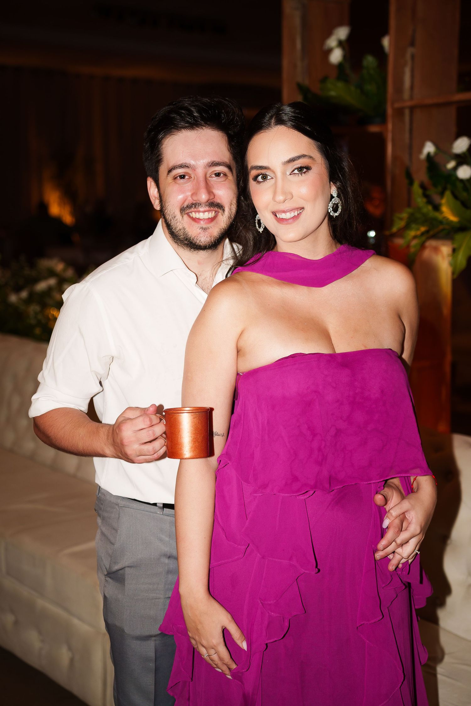 A smiling couple poses at a formal event, man in white shirt holding copper mug, woman in elegant magenta dress.