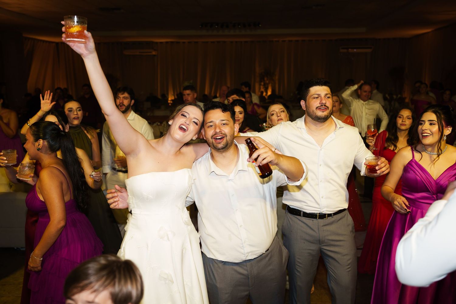 Joyful bride in white dress raises drink with groom and guests celebrating at wedding reception dance floor.