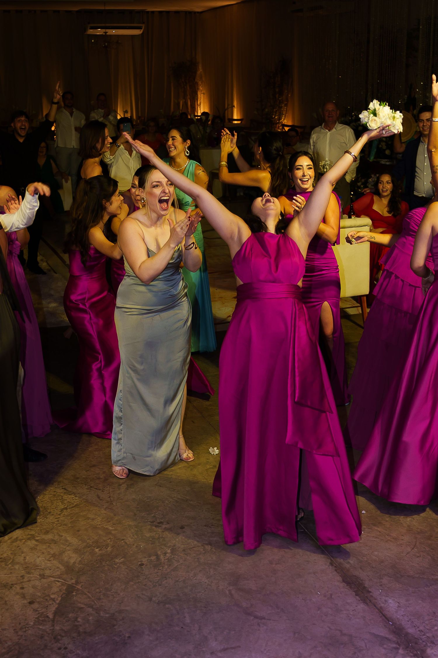 Bridesmaids in magenta gowns dance joyfully at a wedding reception, with one guest in silver reaching for the bouquet toss.