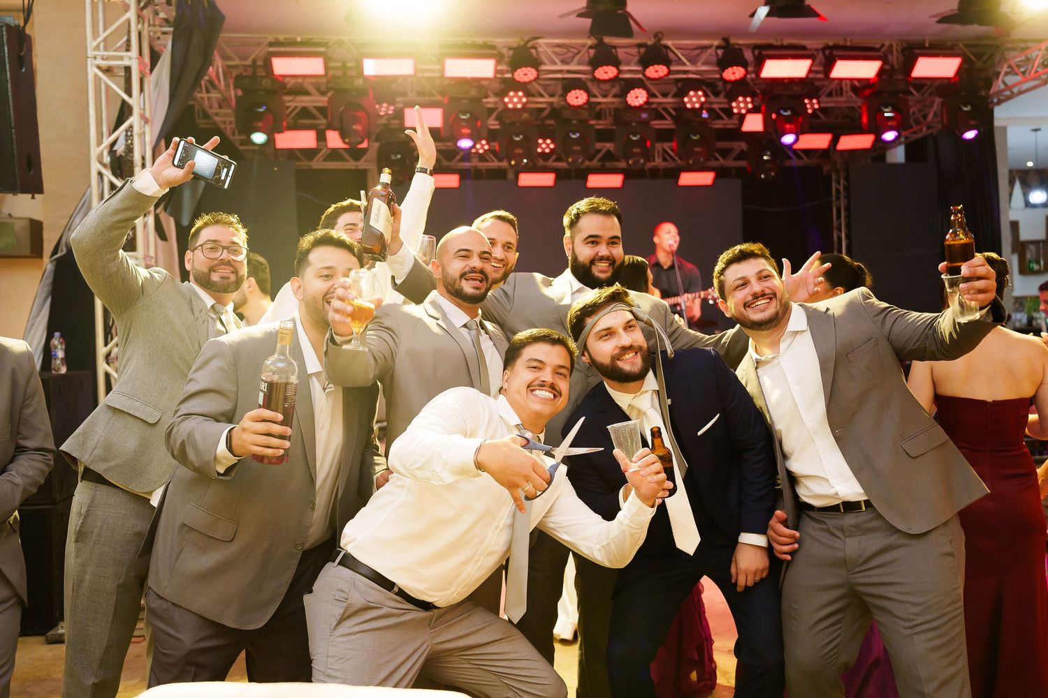 Group of groomsmen celebrating at wedding reception, posing with drinks and big smiles on dance floor.