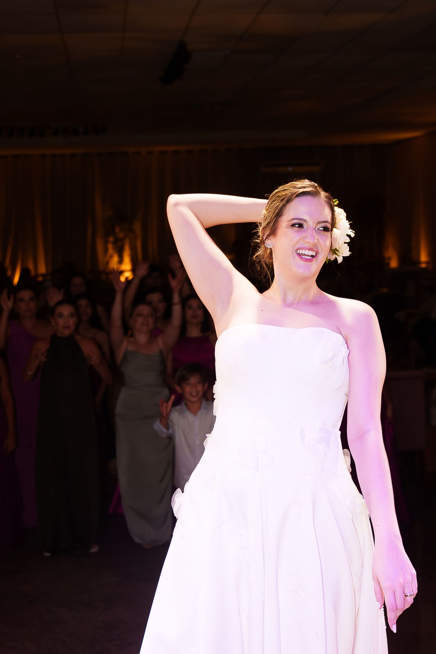 Smiling bride in white strapless gown poses elegantly at wedding reception with guests celebrating in background.