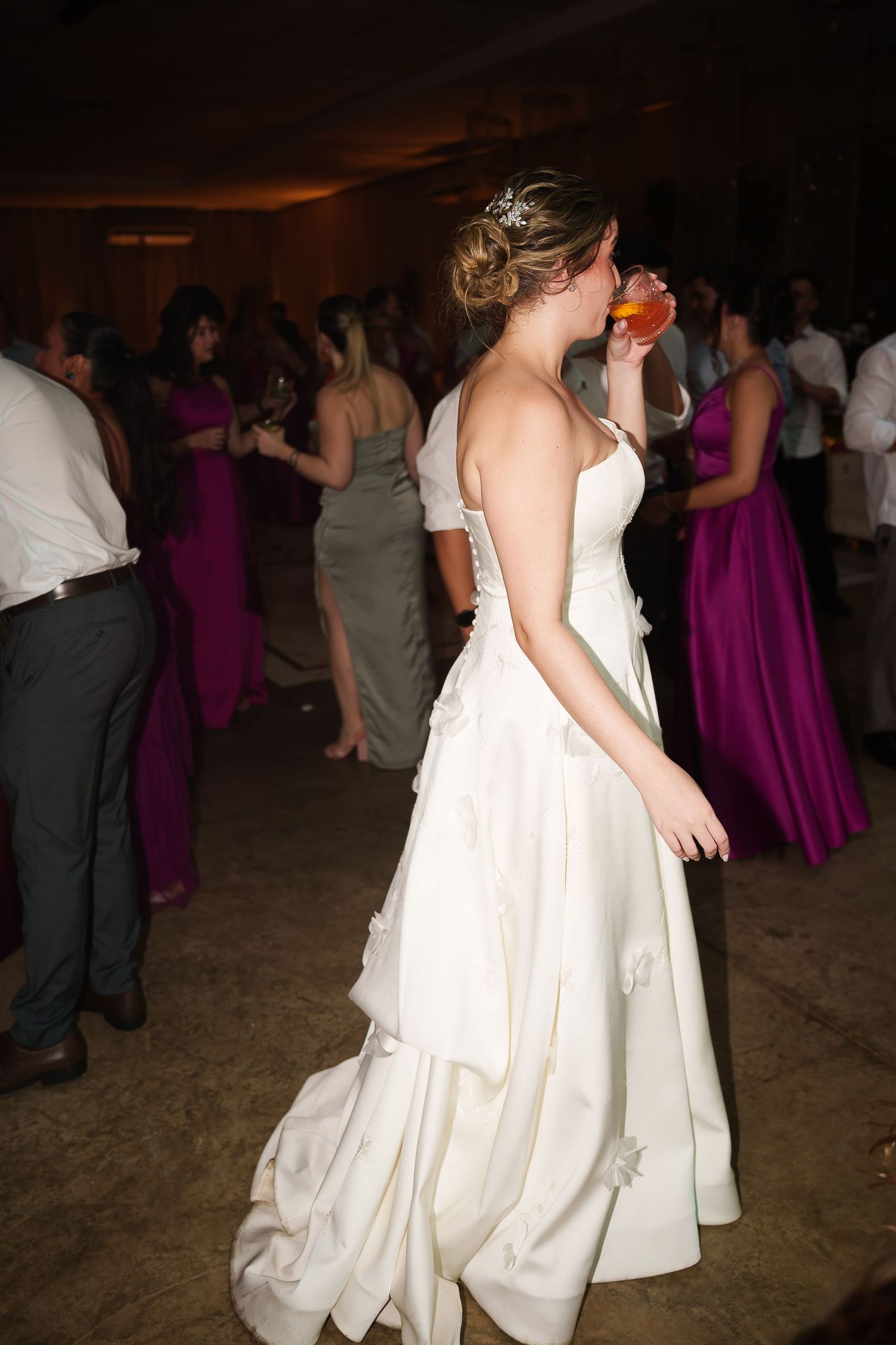 Bride in white gown sipping a drink on the dance floor at a wedding reception with guests in purple dresses.