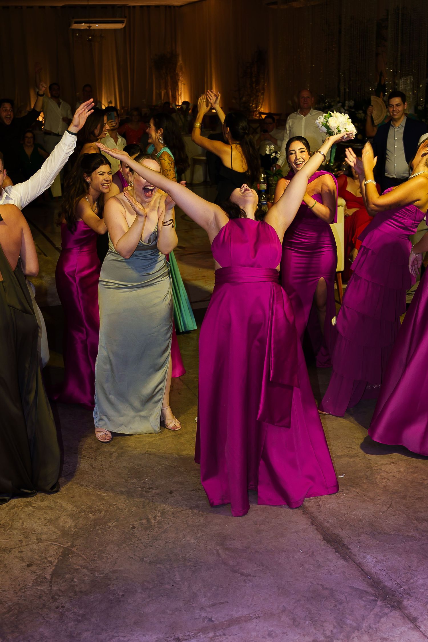 Bridesmaids in magenta and sage gowns dancing joyfully at a wedding reception, reaching for the bouquet toss.