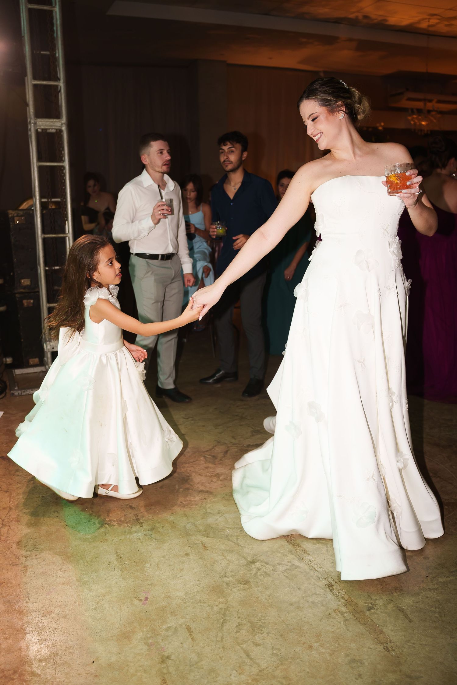 Bride in white strapless gown dancing with flower girl at wedding reception while guests watch in background.