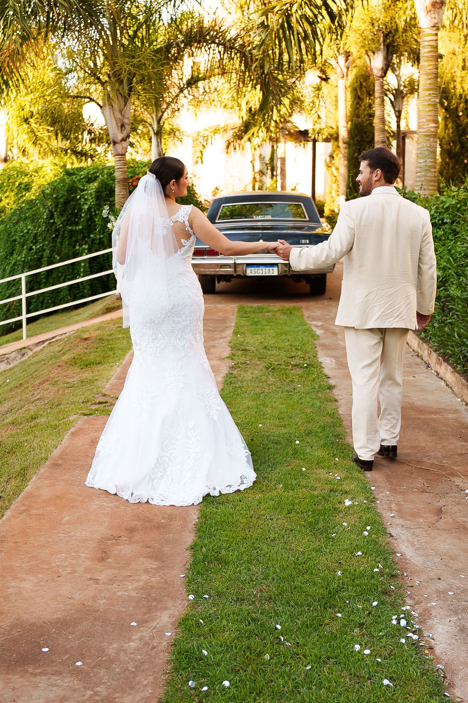 Bride and groom walking hand in hand toward a classic car on a lush garden path on their wedding day.