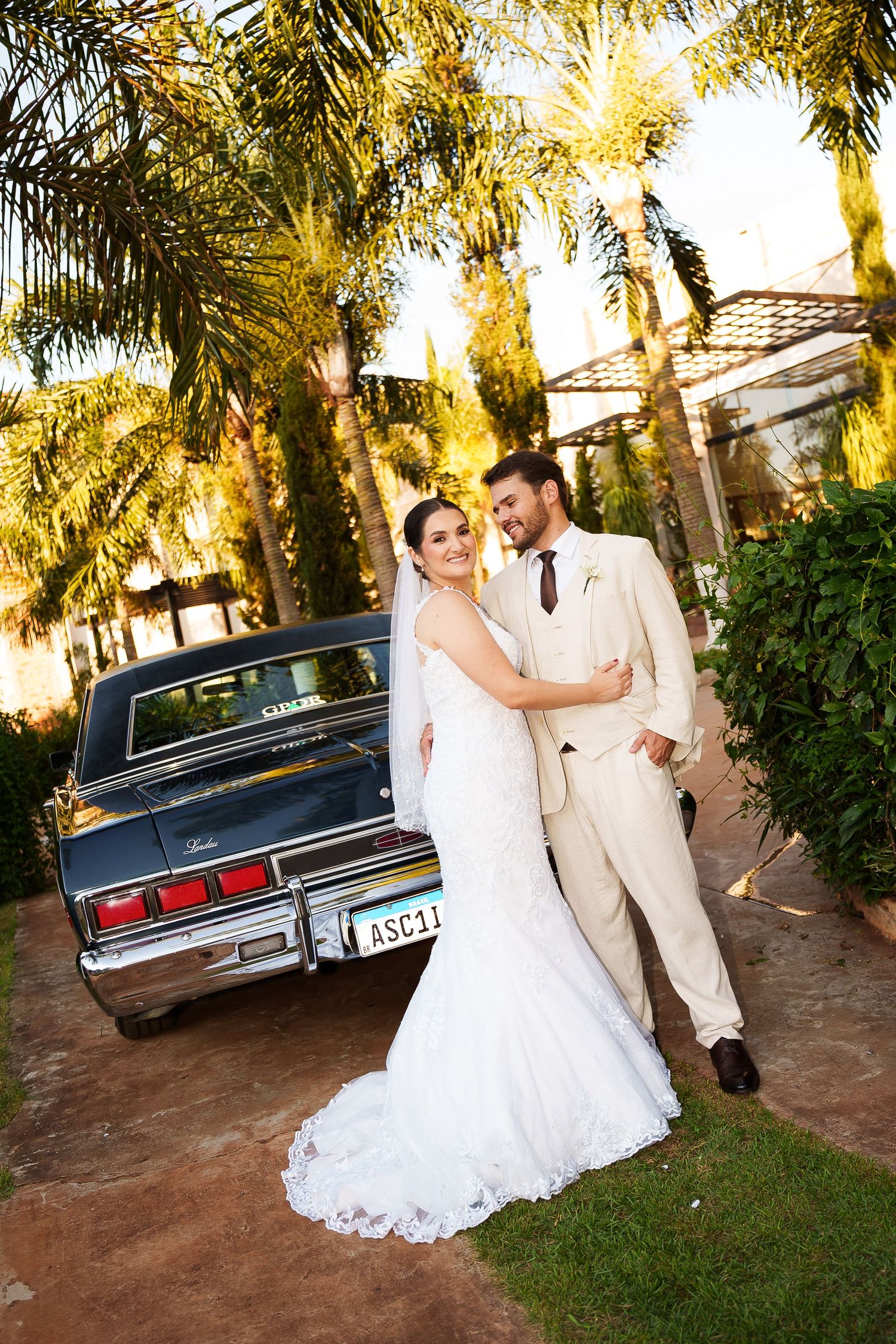 A bride in a white lace gown and groom in a beige suit pose beside a classic car surrounded by palm trees.