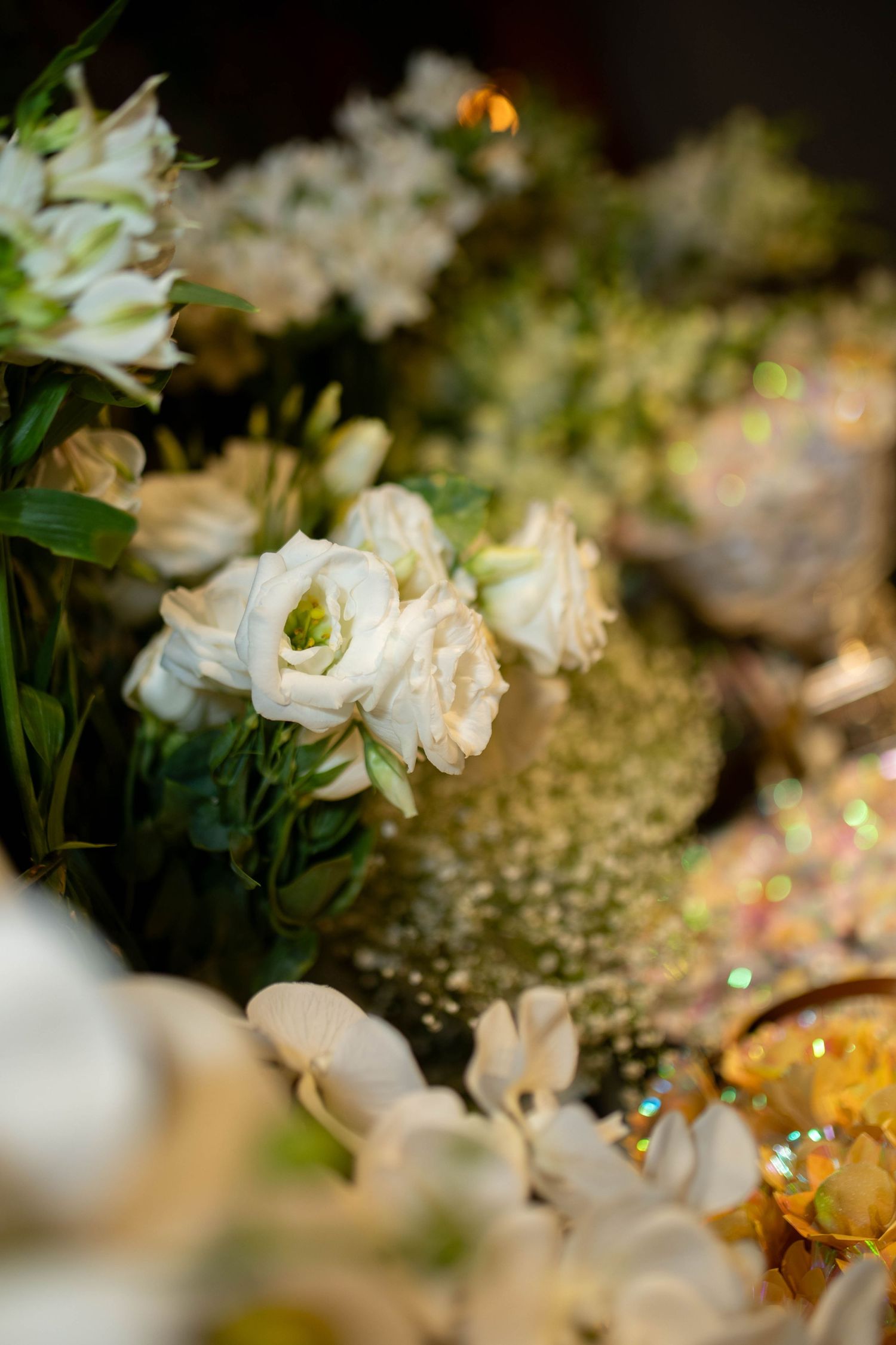 Elegant white roses and lisianthus flowers with green foliage and golden glitter accents in a wedding table arrangement.