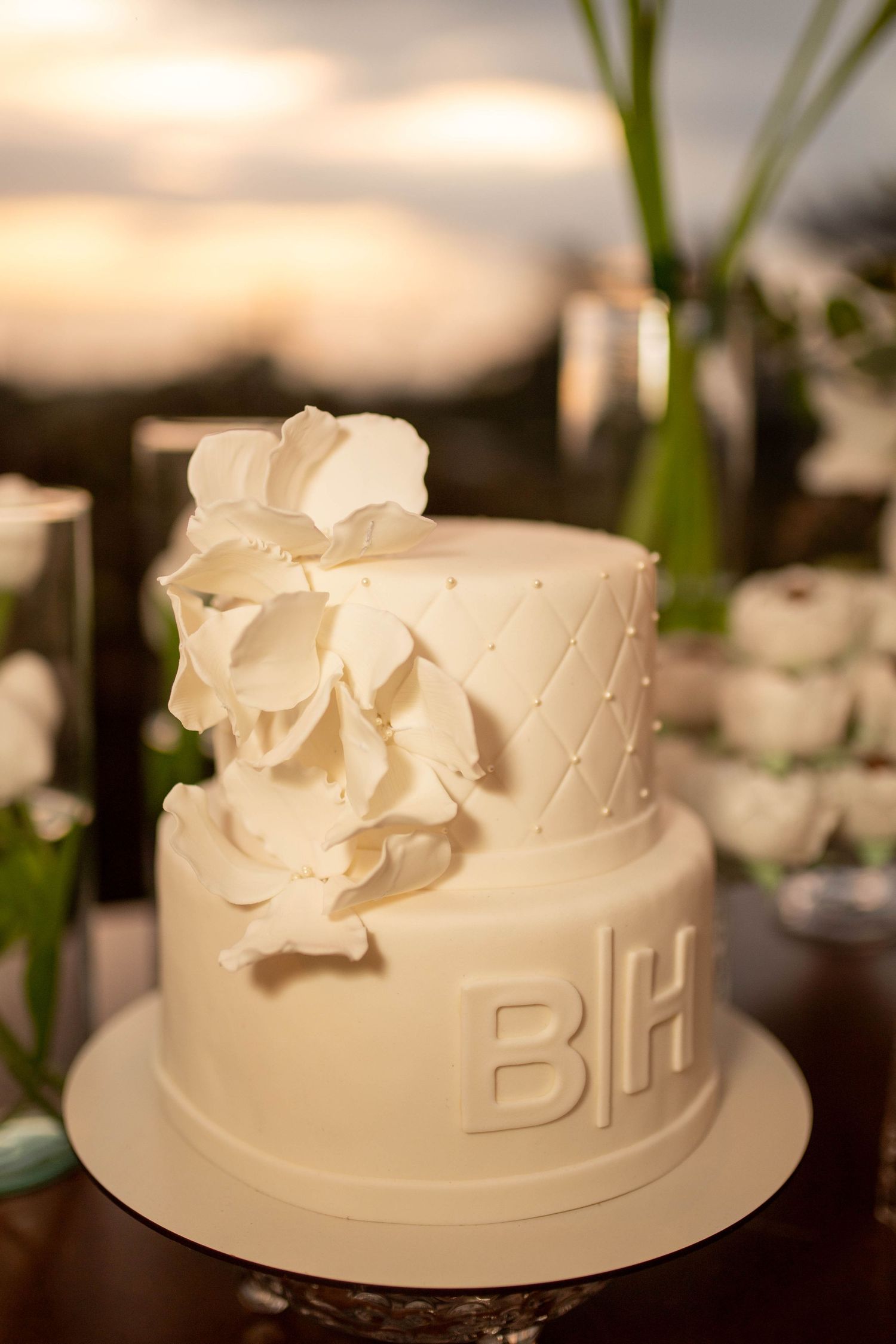 Elegant two-tier white fondant wedding cake with monogram BH and floral decorations at sunset.