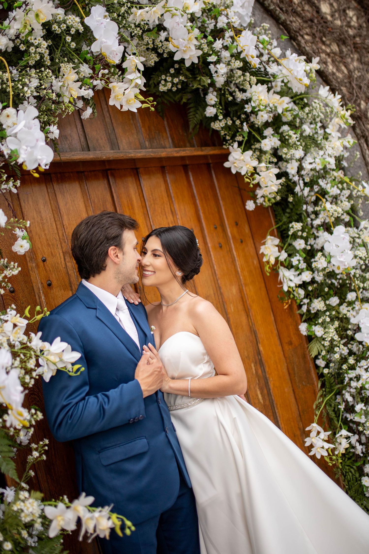 Bride and groom embrace under a floral arch with white flowers and a wooden door backdrop.