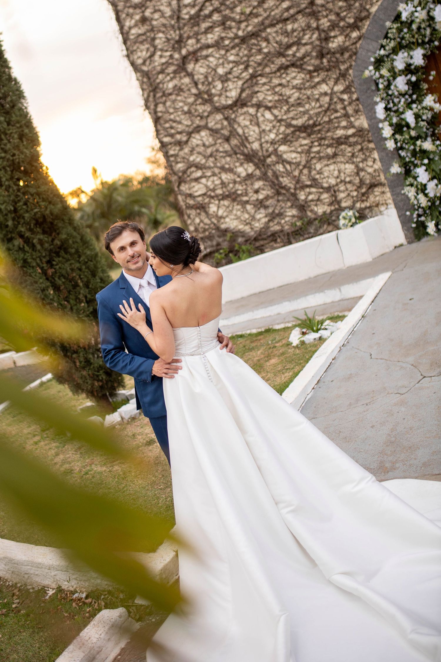 A bride in a white strapless gown and groom in blue suit share a romantic embrace outdoors at golden hour.
